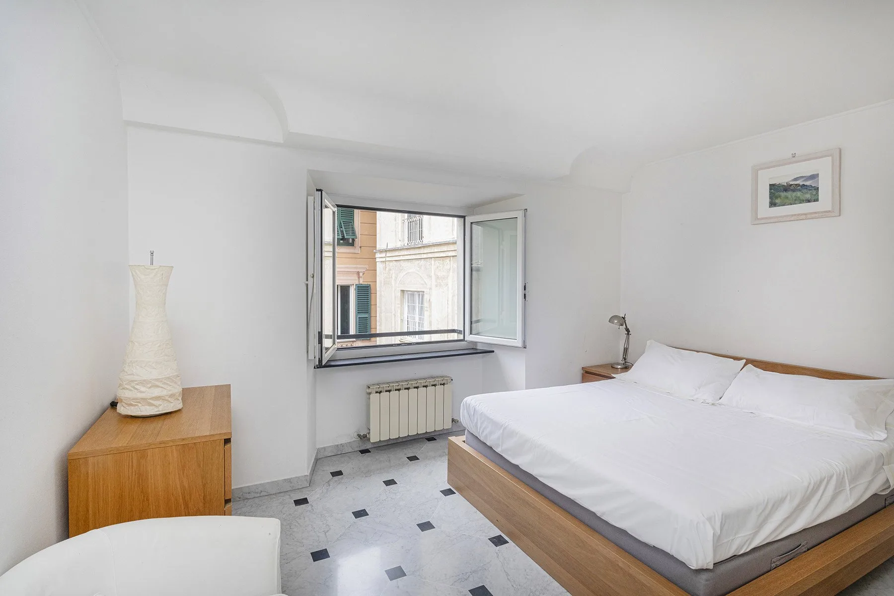Un dormitorio luminoso con una cama, una cómoda de madera y una ventana abierta.