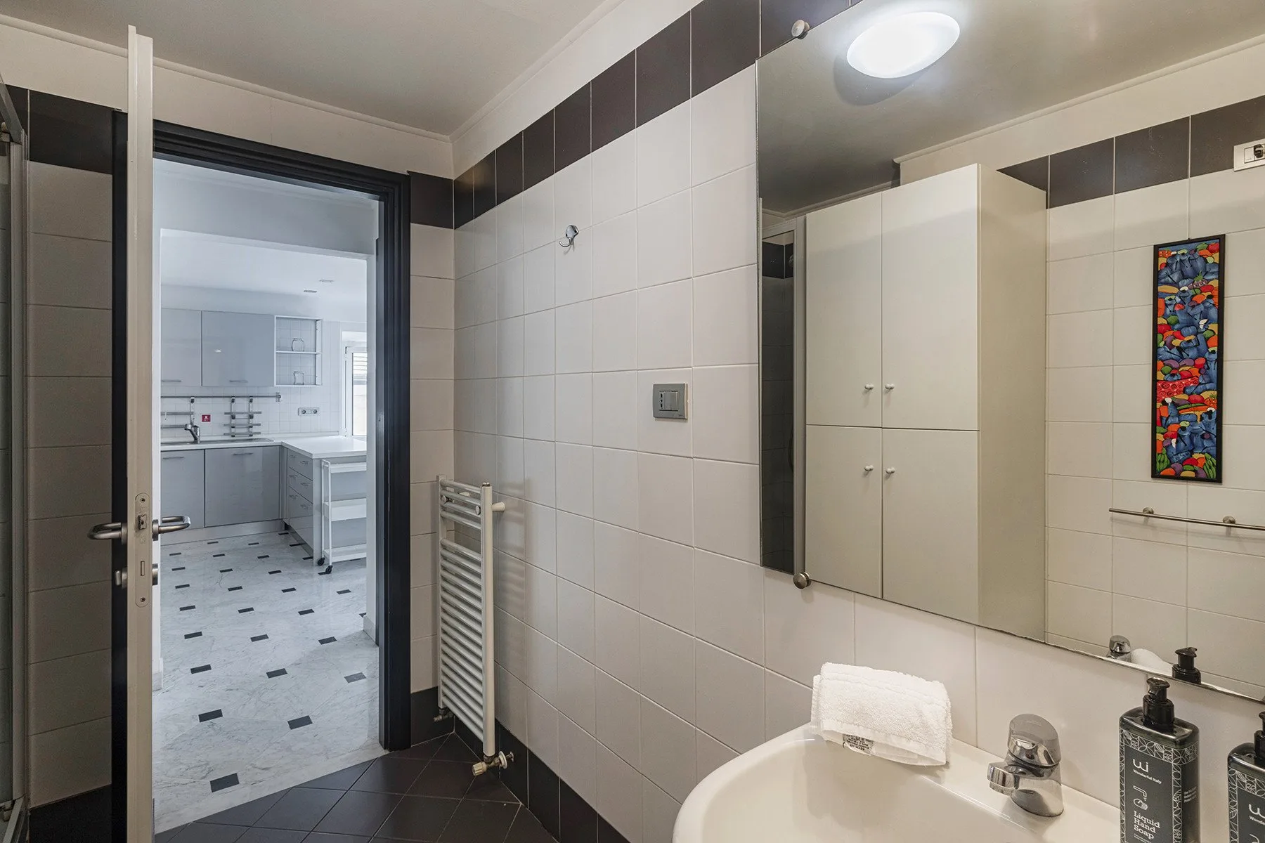 Un baño con azulejos, lavabo y una vista de la cocina a través de la puerta.