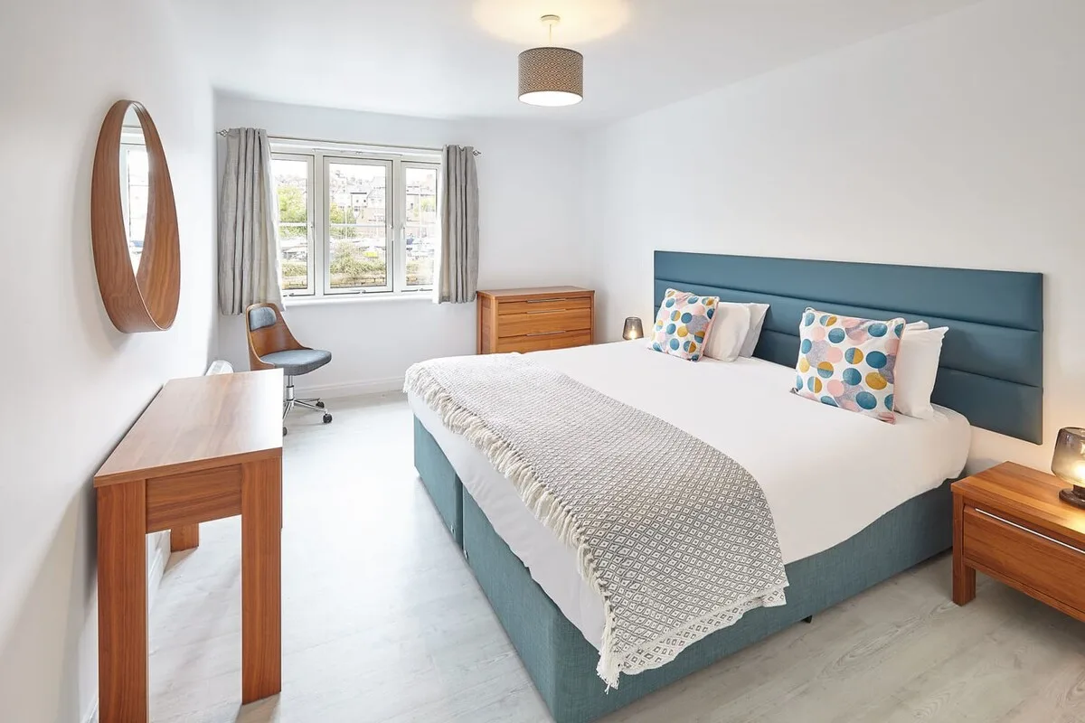 A bright bedroom with a bed, wooden furnishings, and a window with curtains.