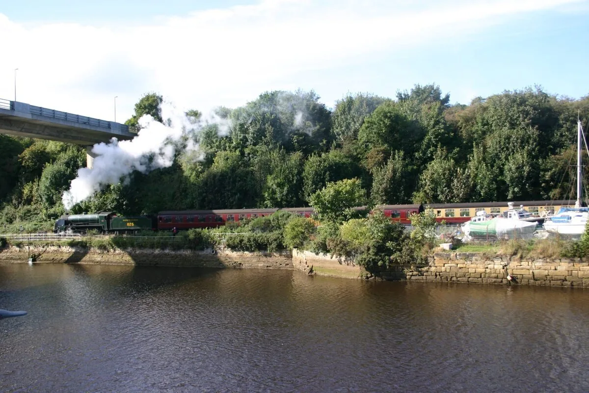A steam train travels beside a river, with boats and trees along the bank.