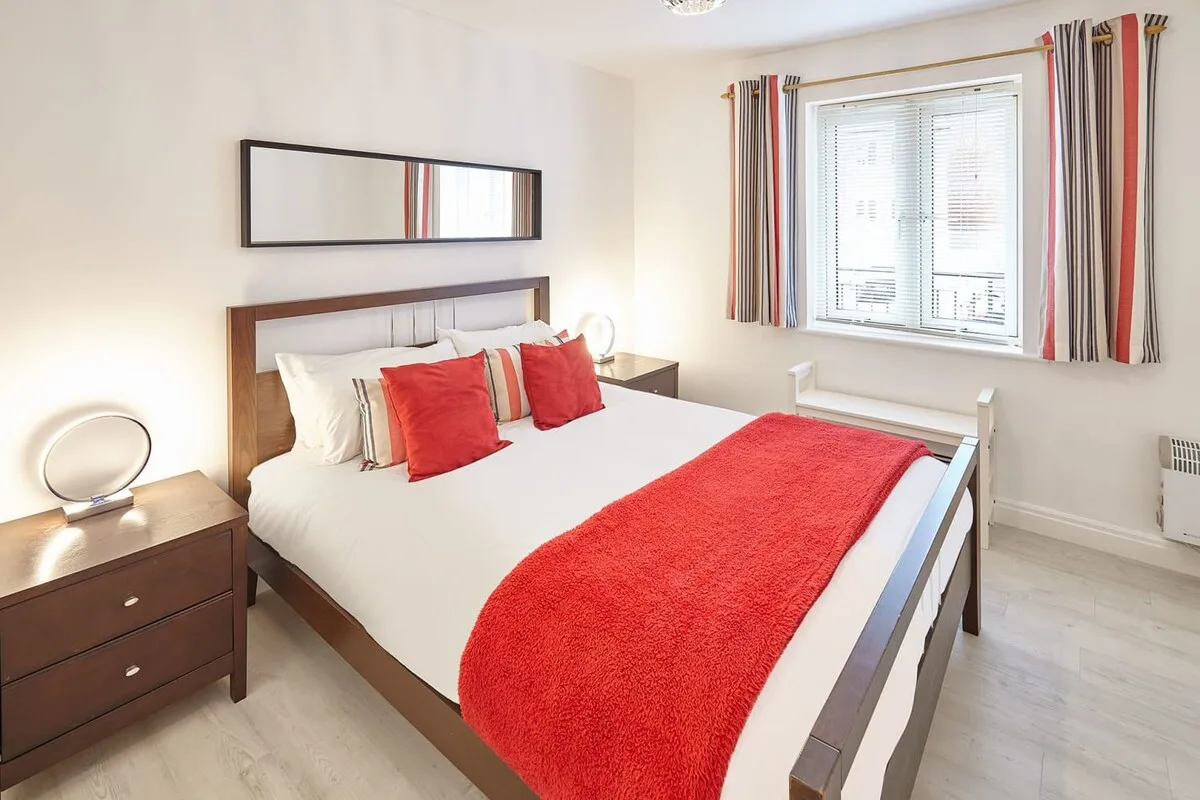 A bedroom with a double bed dressed in white and red bedding beside a window.