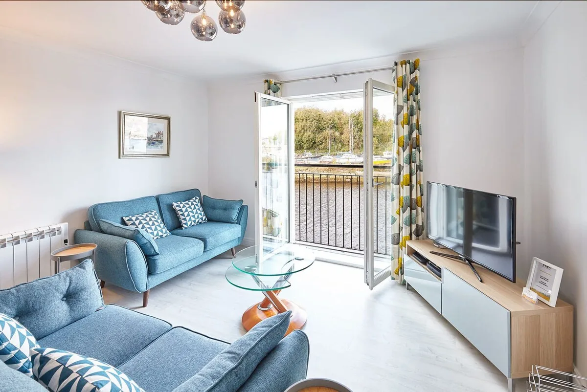 A bright living room with blue seating, a glass coffee table, and open balcony doors.