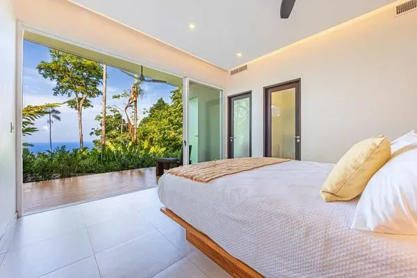 Dormitorio luminoso con una cama junto a grandes ventanales con vistas a la vegetación y al océano.