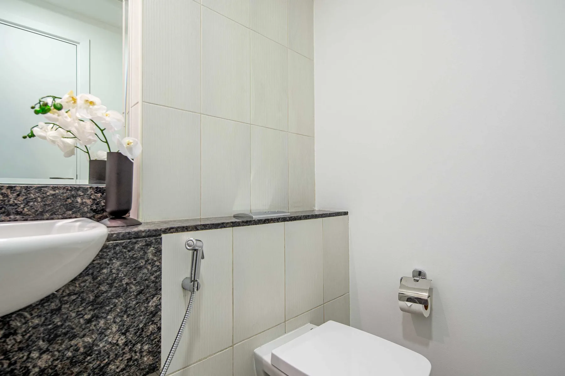 Un baño alicatado con lavabo, inodoro y ducha higiénica montada en la pared.