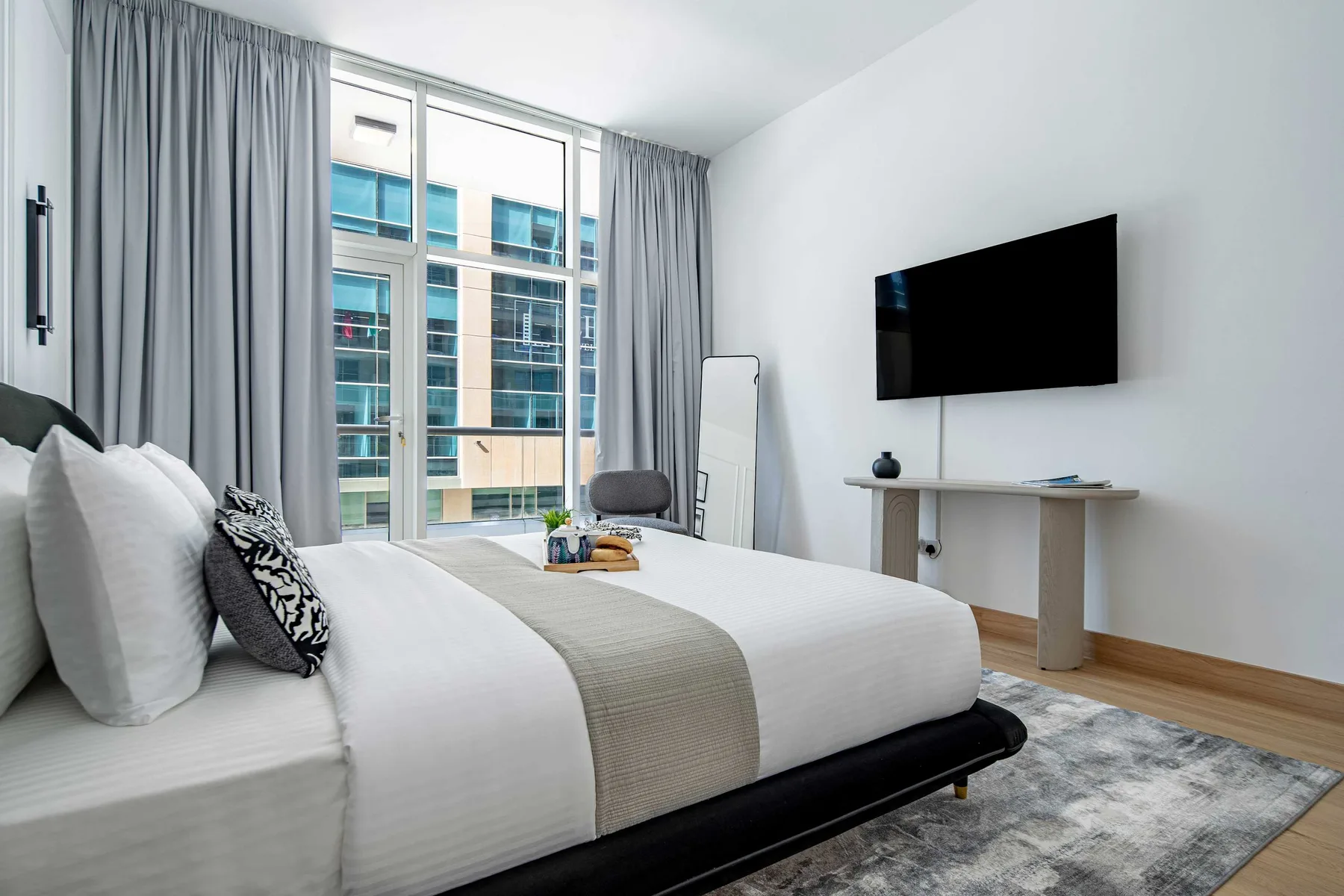 Un dormitorio luminoso con una cama tendida, cortinas grises y un televisor en la pared.