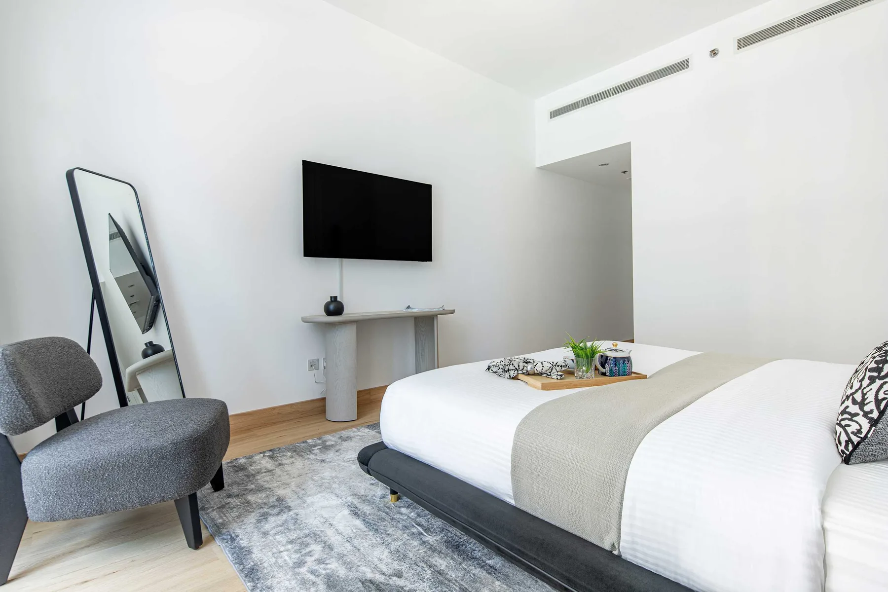 Un dormitorio luminoso con una cama tendida, un televisor en la pared y asientos grises.