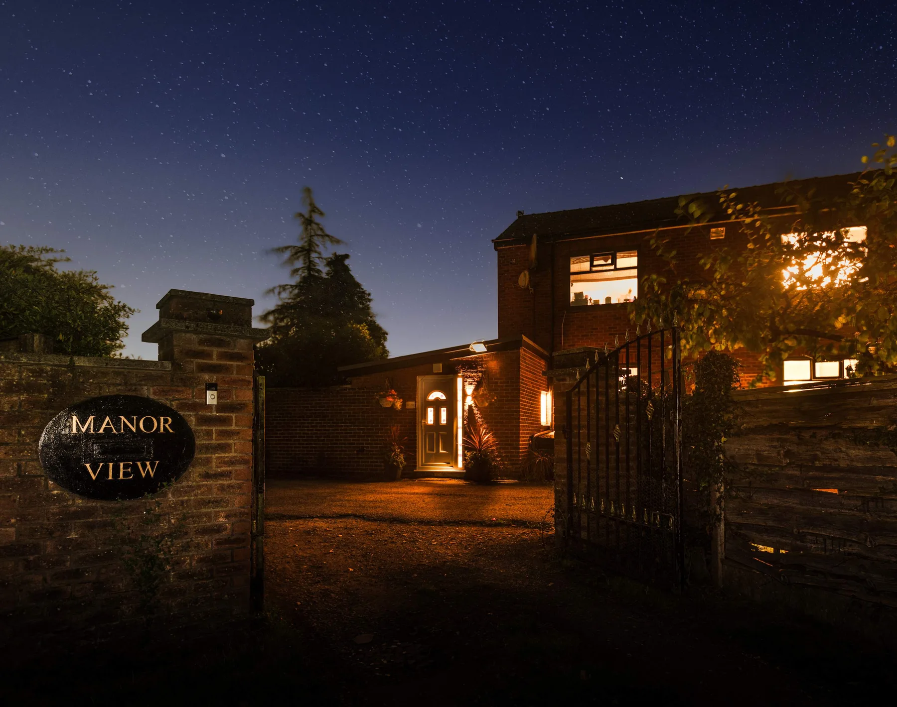 A brick manor entrance and open gate are illuminated beneath a starry night sky.