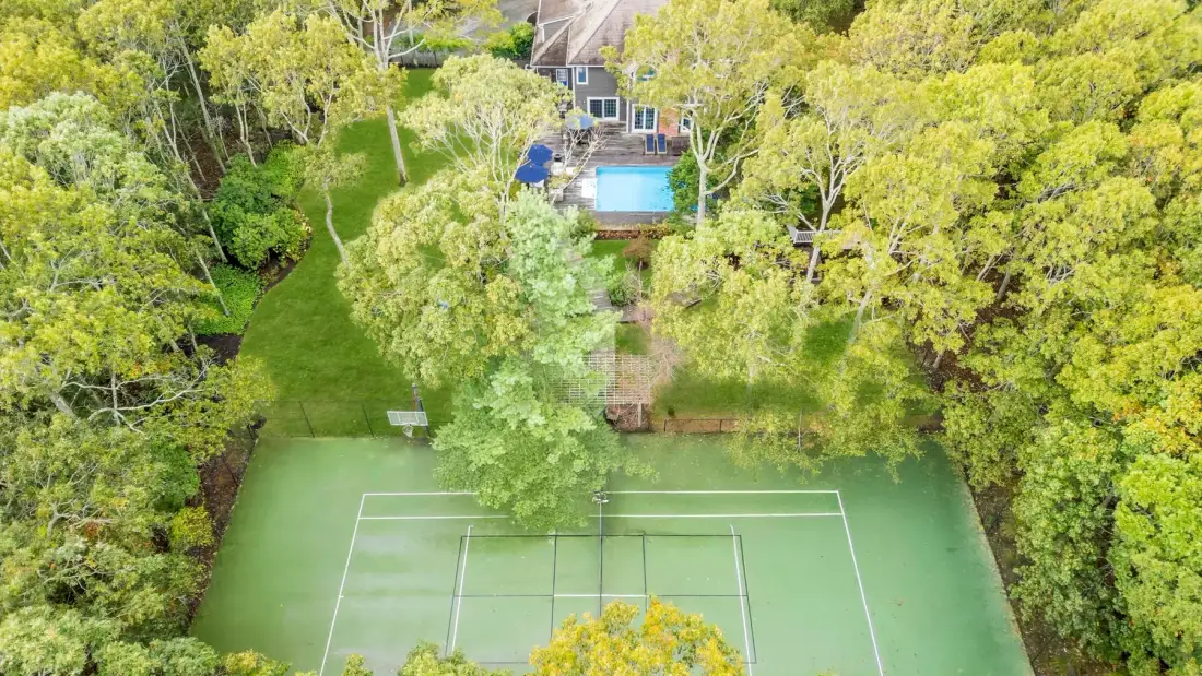 Vista aérea de una cancha de tenis rodeada de árboles, con una piscina al fondo.