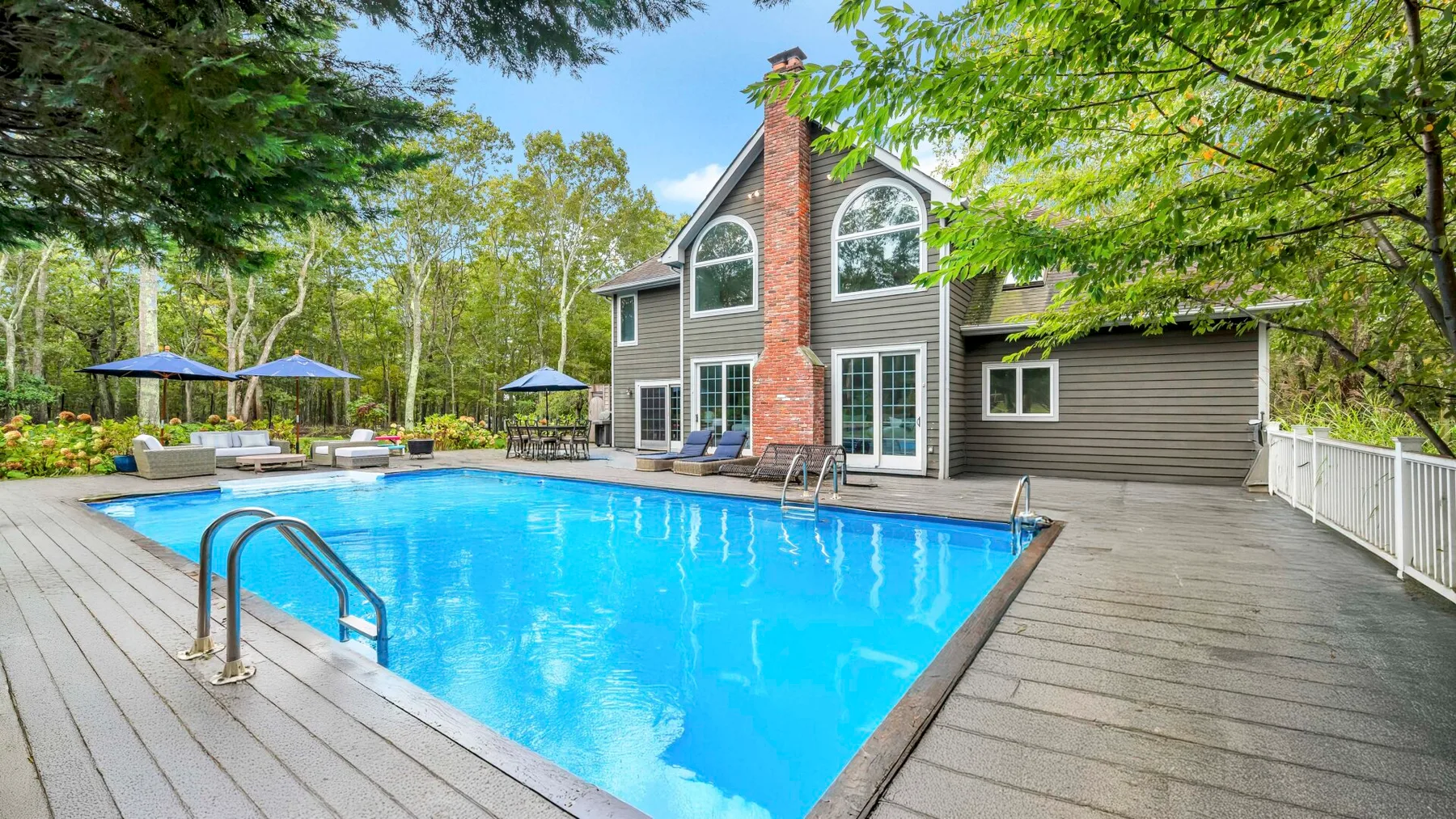 Una piscina al aire libre junto a una casa, rodeada de una terraza de madera y árboles.