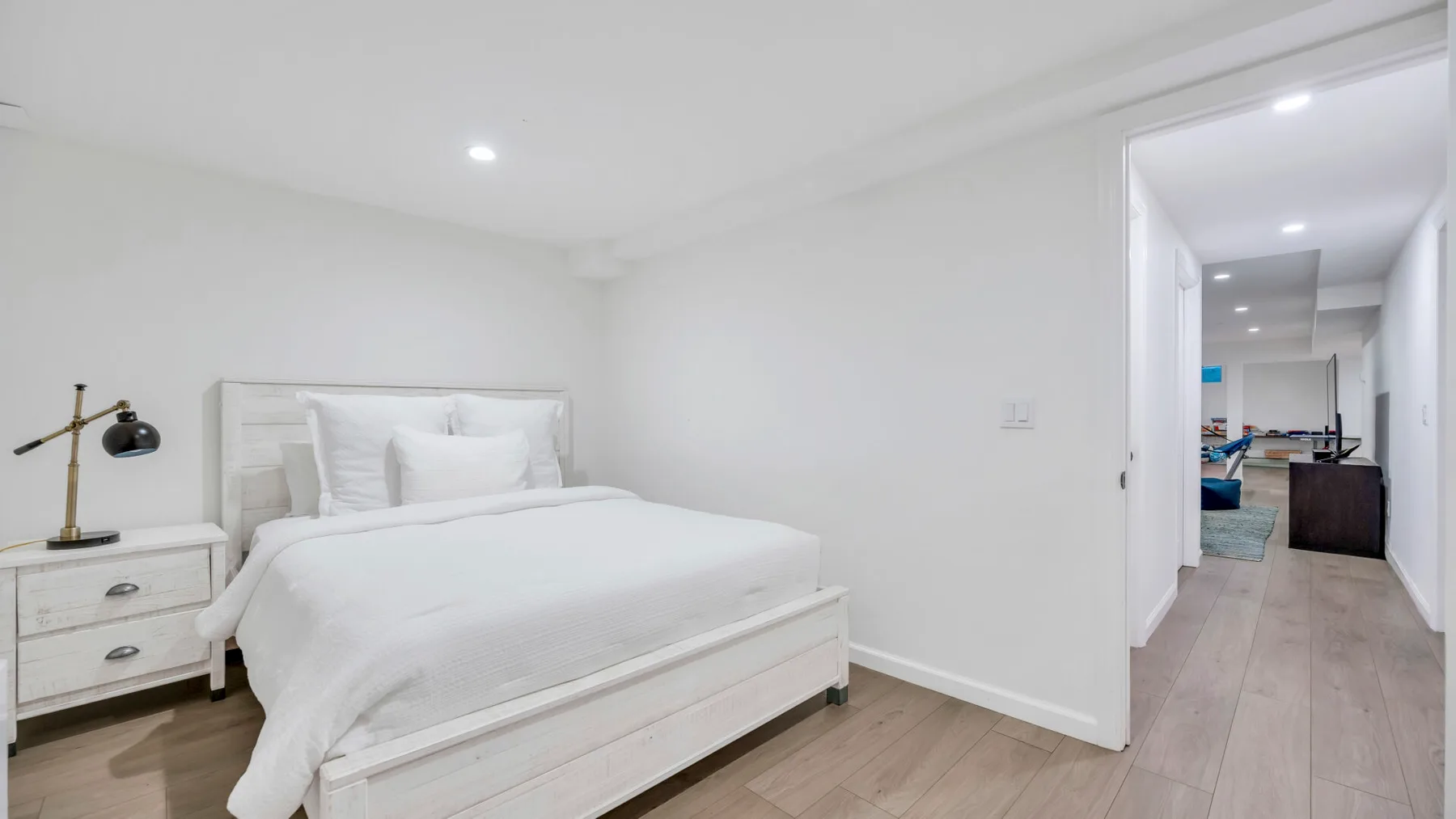 Un dormitorio luminoso con una cama blanca y una puerta abierta al pasillo.
