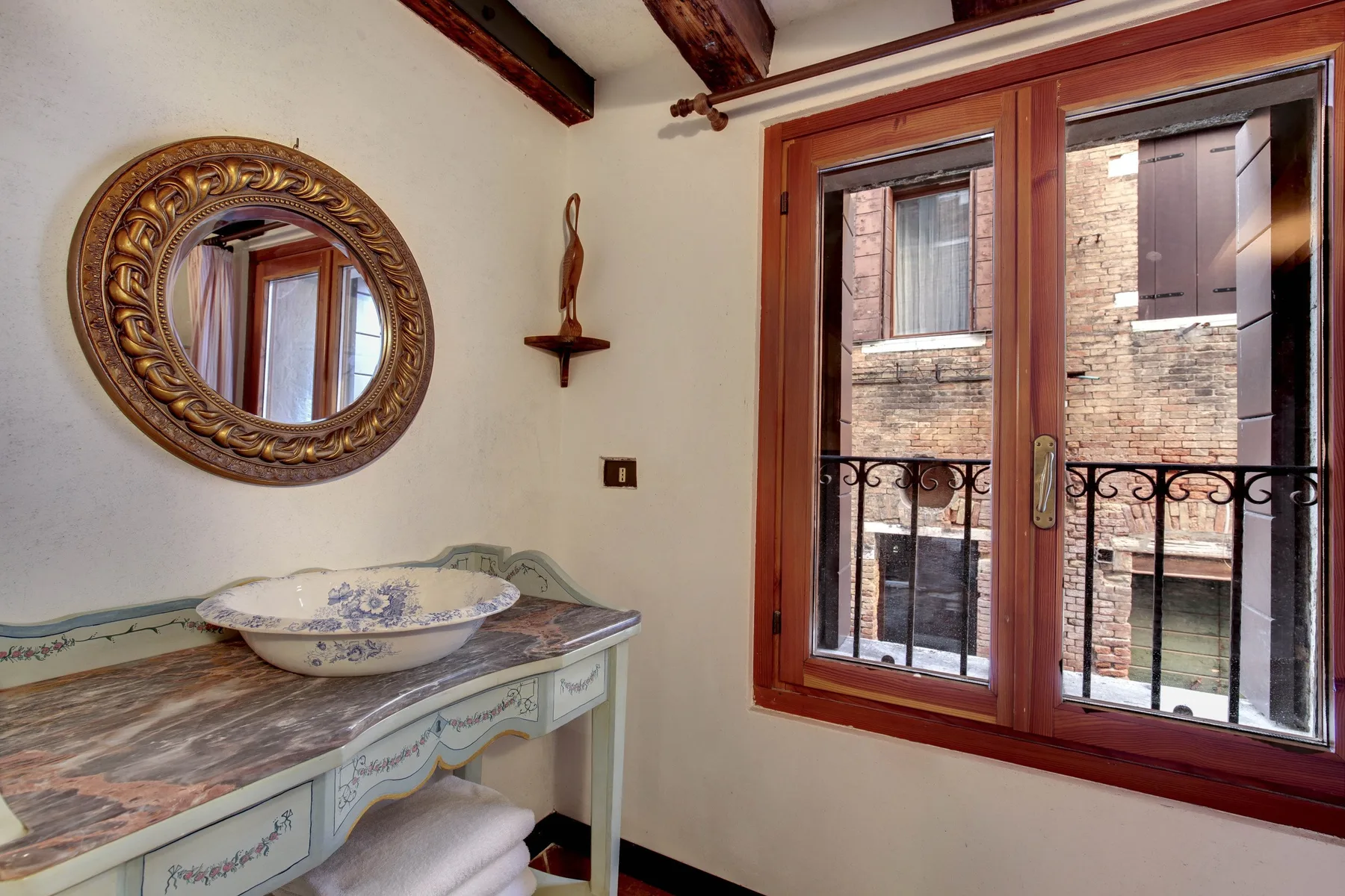 Un tocador de baño con un espejo decorativo está junto a una ventana con marco de madera.