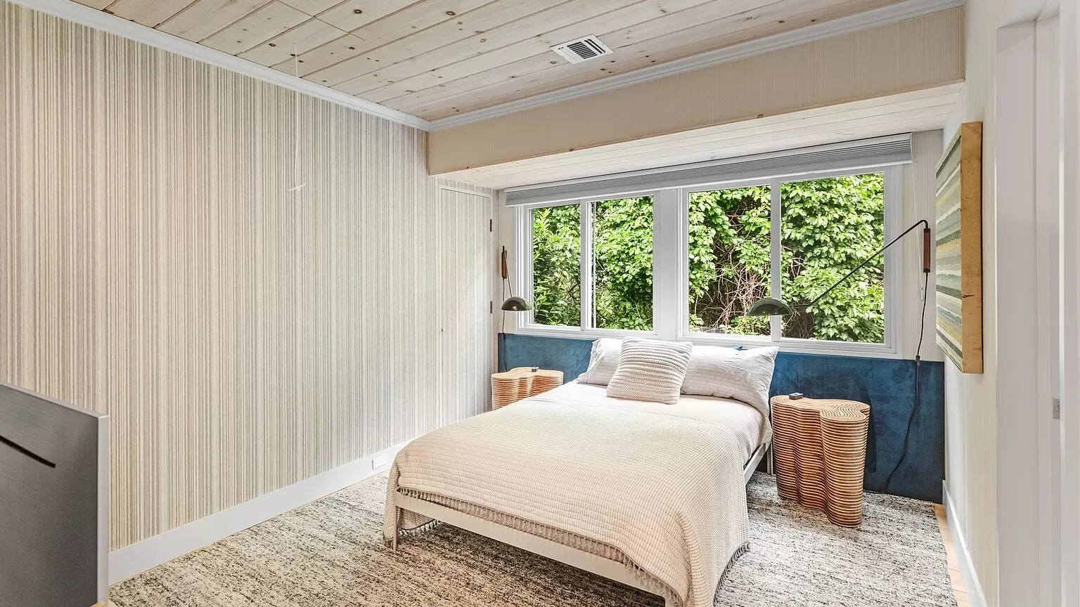 Un dormitorio con una cama bajo una ventana amplia con vegetación a la vista.