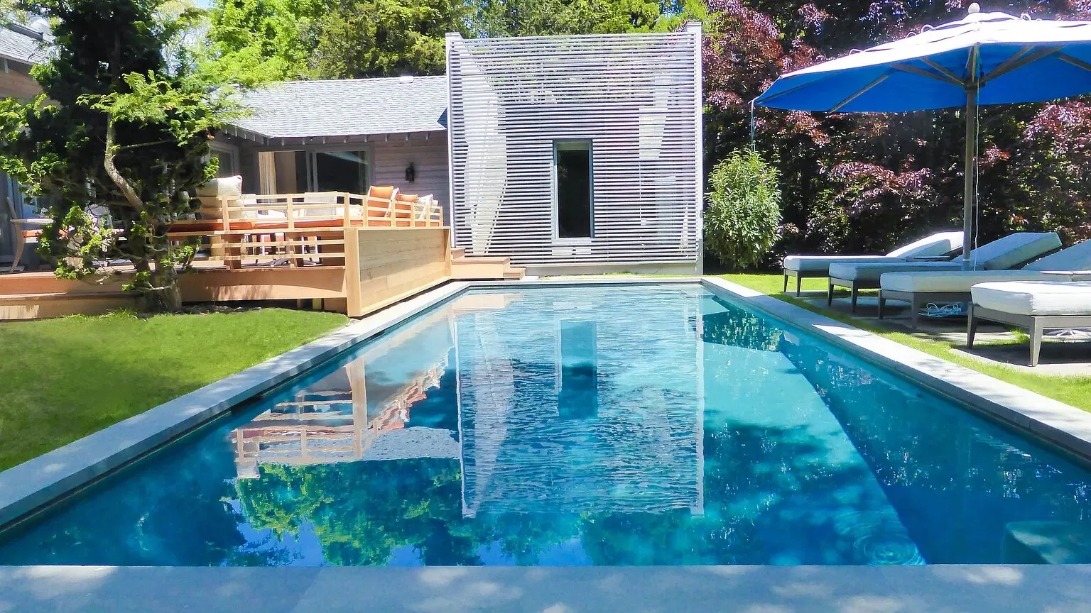 Una piscina al aire libre junto a una terraza, con asientos reclinables y una sombrilla azul.