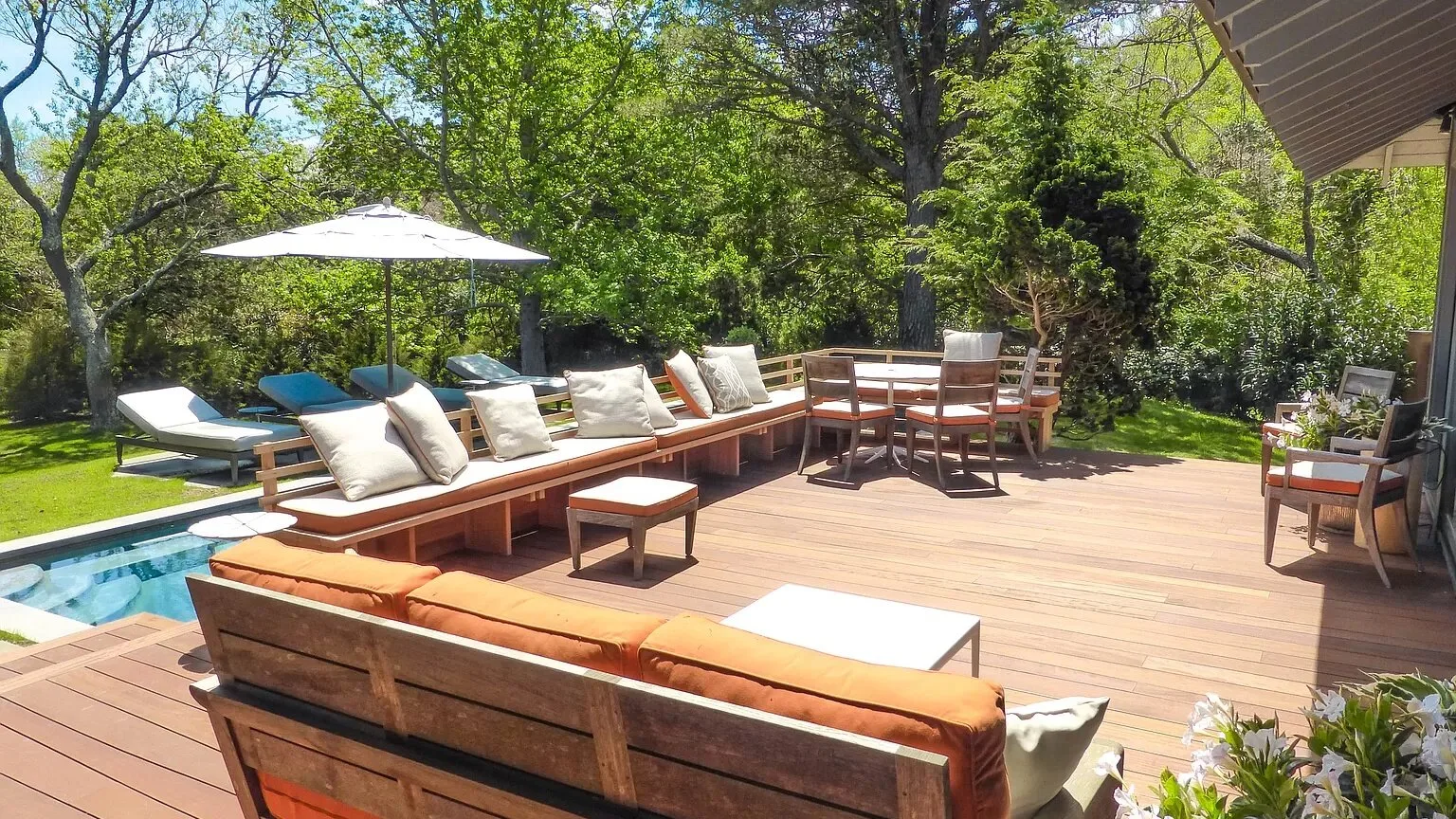 Una terraza exterior soleada tiene asientos acolchados alrededor de una mesa pequeña junto a un jardín.