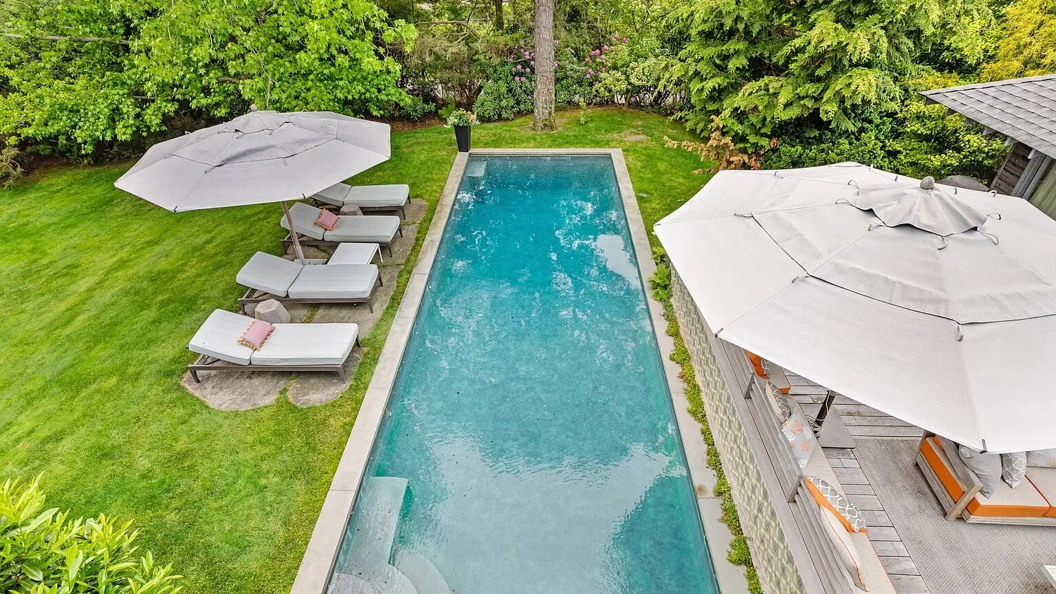Una piscina rectangular al aire libre está rodeada de césped, vegetación y asientos a la sombra.