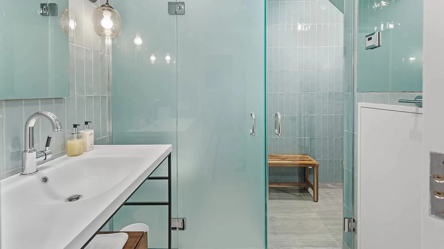 Un baño con azulejos, un largo lavabo y una mampara de ducha de vidrio.