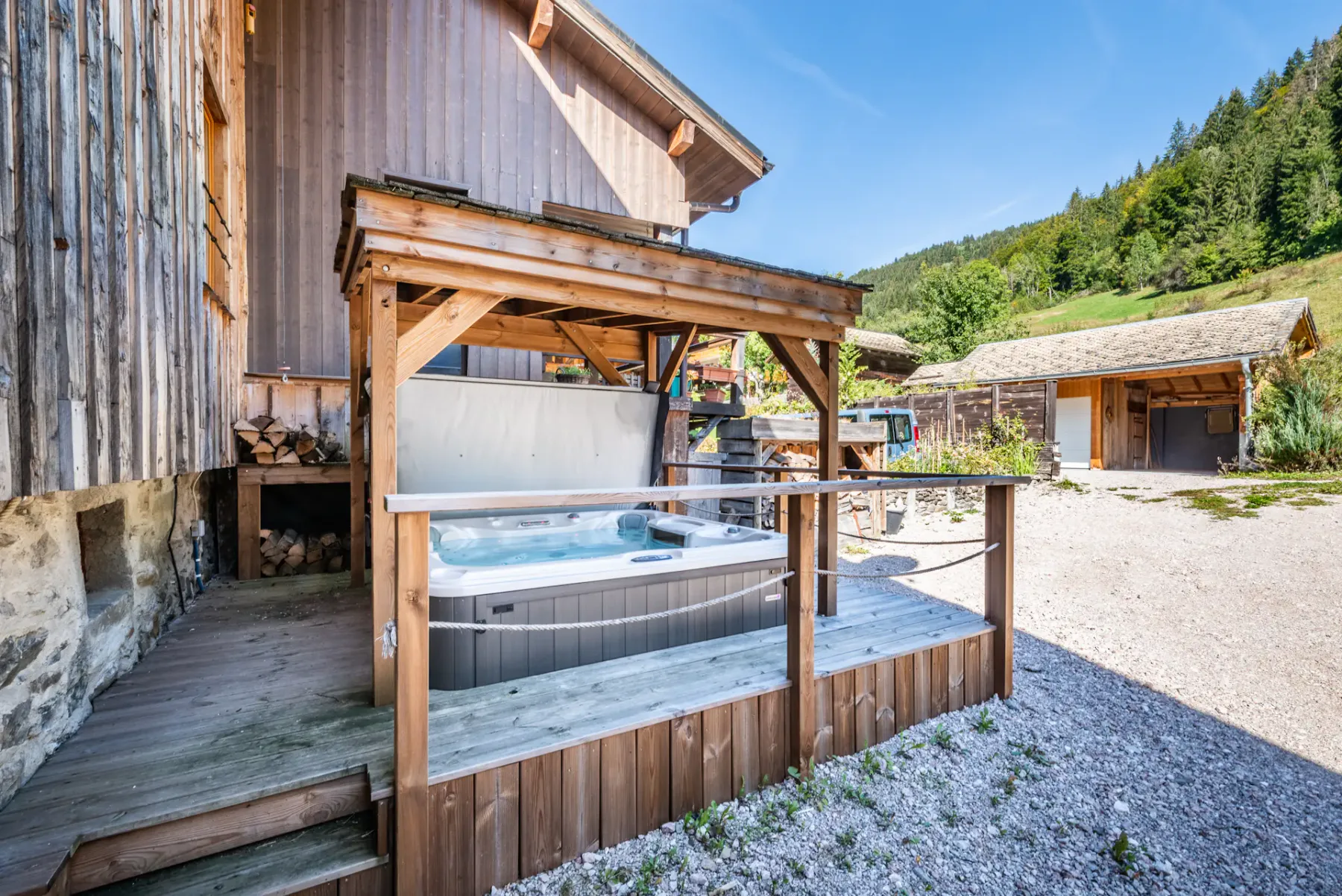 An outdoor hot tub sits beside a wooden chalet, with a forested hillside behind it.