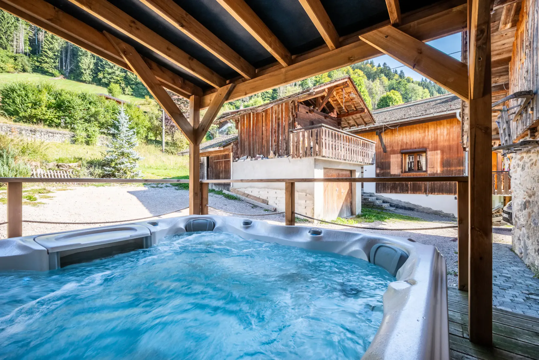 An outdoor hot tub sits beneath a wooden shelter, with chalet buildings and green hills beyond.