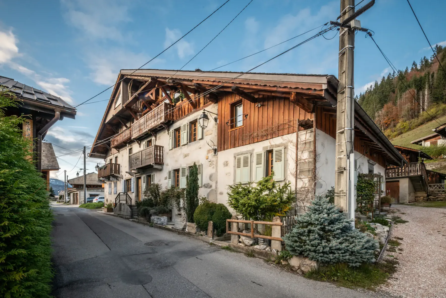 A chalet-style building stands beside a narrow village street with trees and mountain slopes nearby.