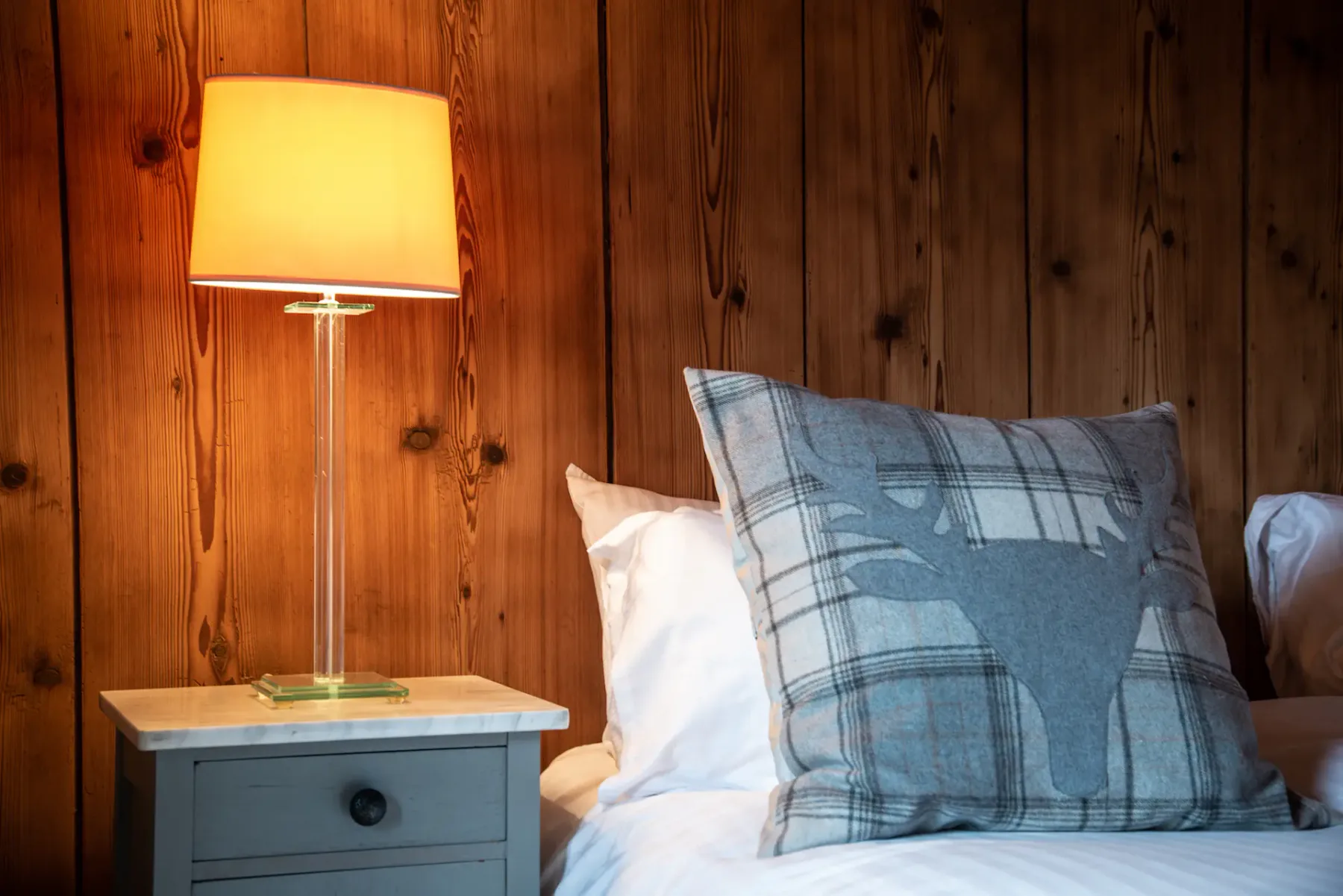 A bedroom with a bed, plaid pillow, and bedside lamp against wood-paneled walls.