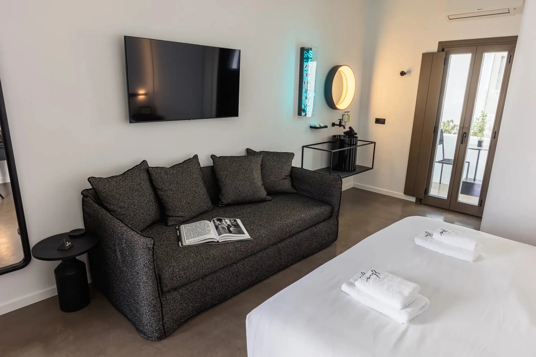 Una zona de estar en un dormitorio, con un sofá oscuro, un televisor en la pared y una cama en primer plano.