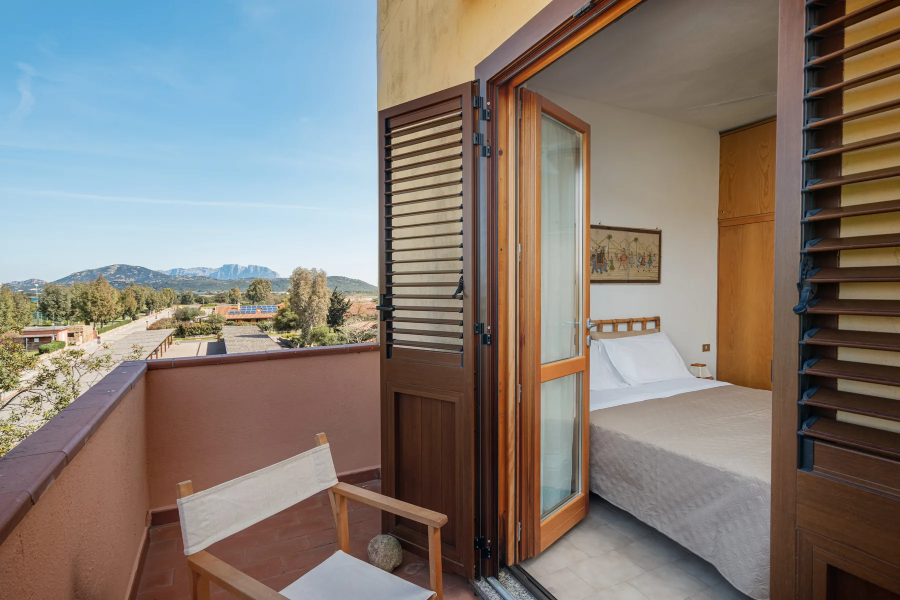 A bedroom opens onto a balcony with a wooden chair and distant hills.