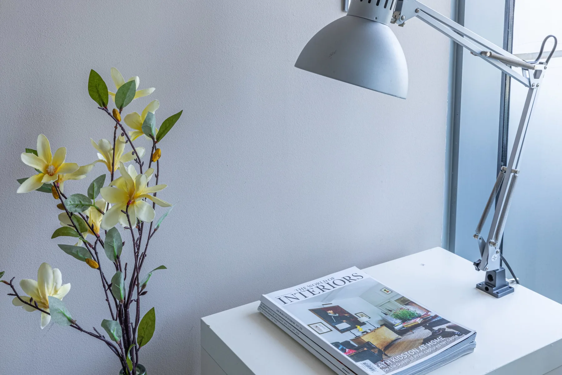 Un escritorio blanco sostiene una revista bajo una lámpara ajustable, con flores al lado.