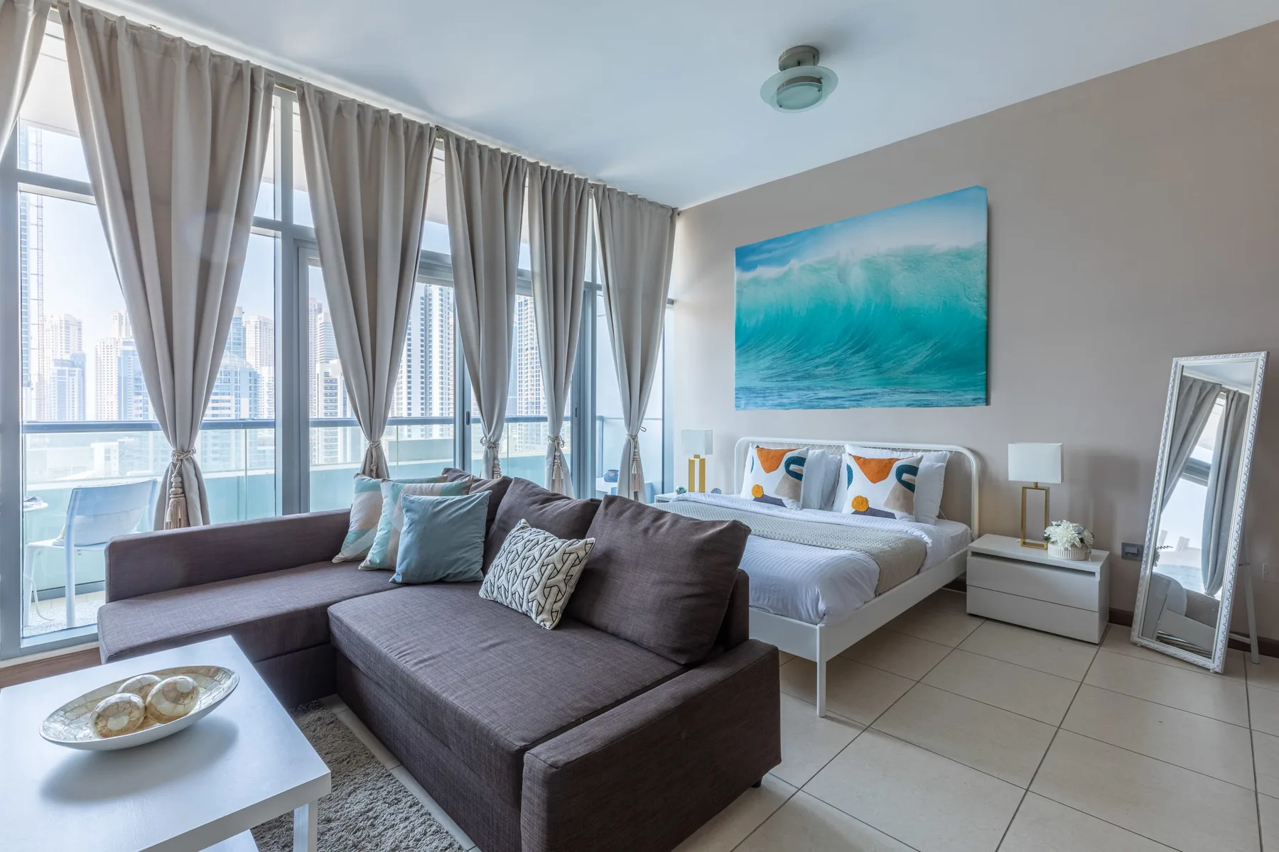 Un dormitorio luminoso con cama, zona de estar y ventanales con edificios de la ciudad.