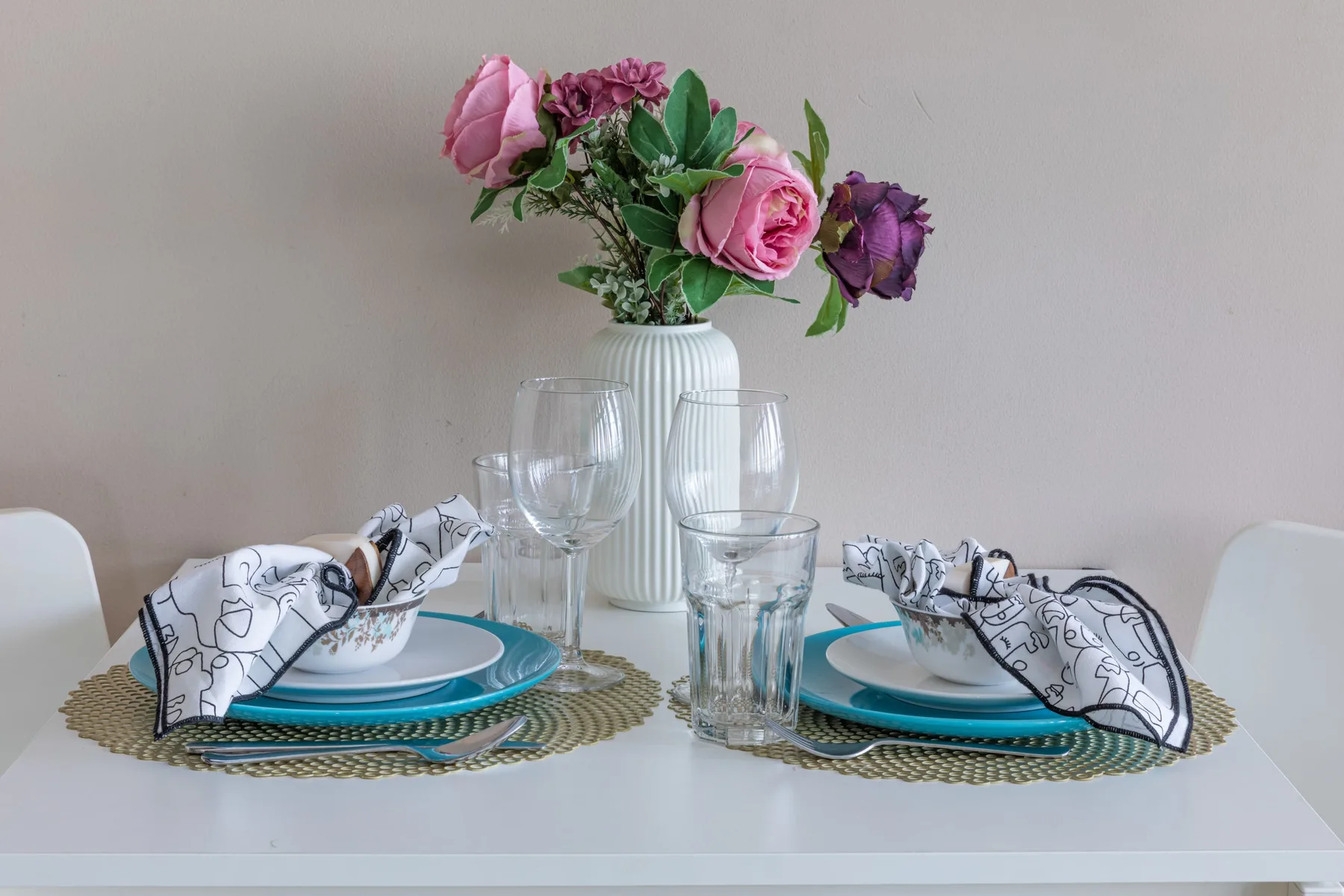 Una mesa de comedor está preparada con platos, vasos, servilletas dobladas y un jarrón de flores.