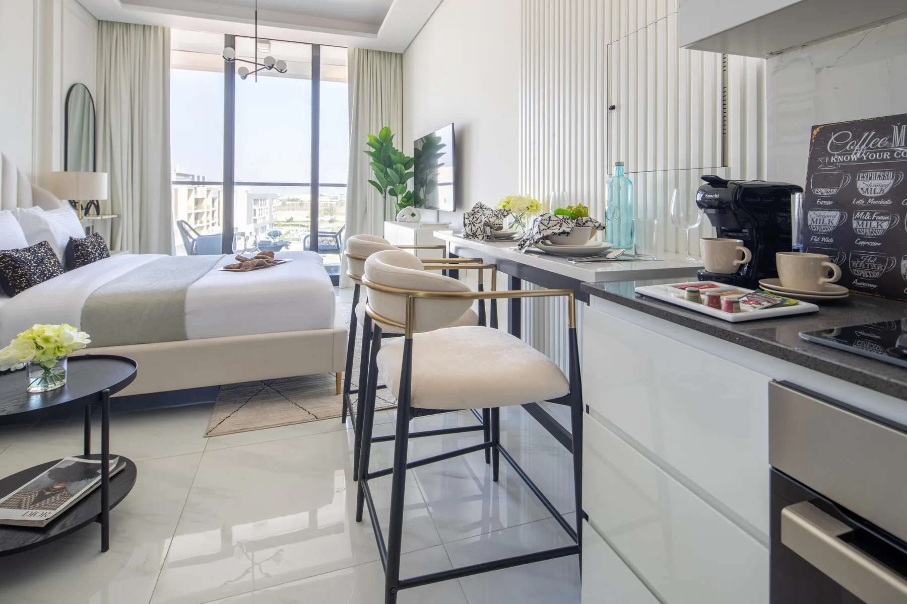 A bright bedroom with a bed, seating area, and kitchenette beside large windows.