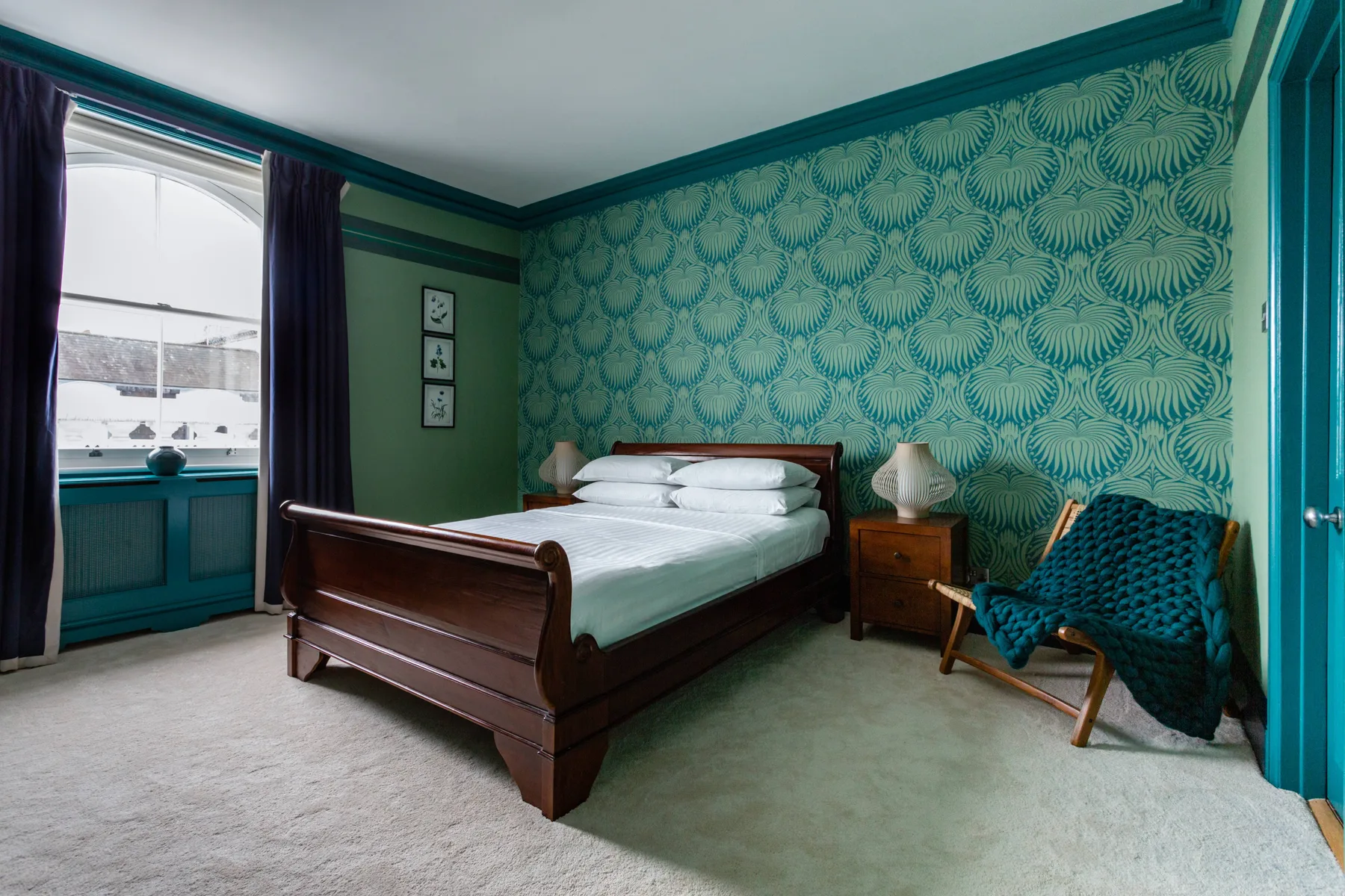 A bedroom with a wooden bed, patterned teal wallpaper, and a window with dark curtains.