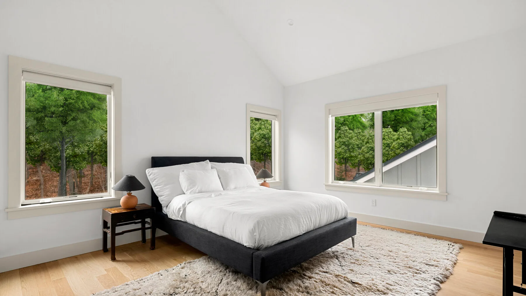 Un dormitorio luminoso con una cama junto a grandes ventanas que dan a árboles verdes.