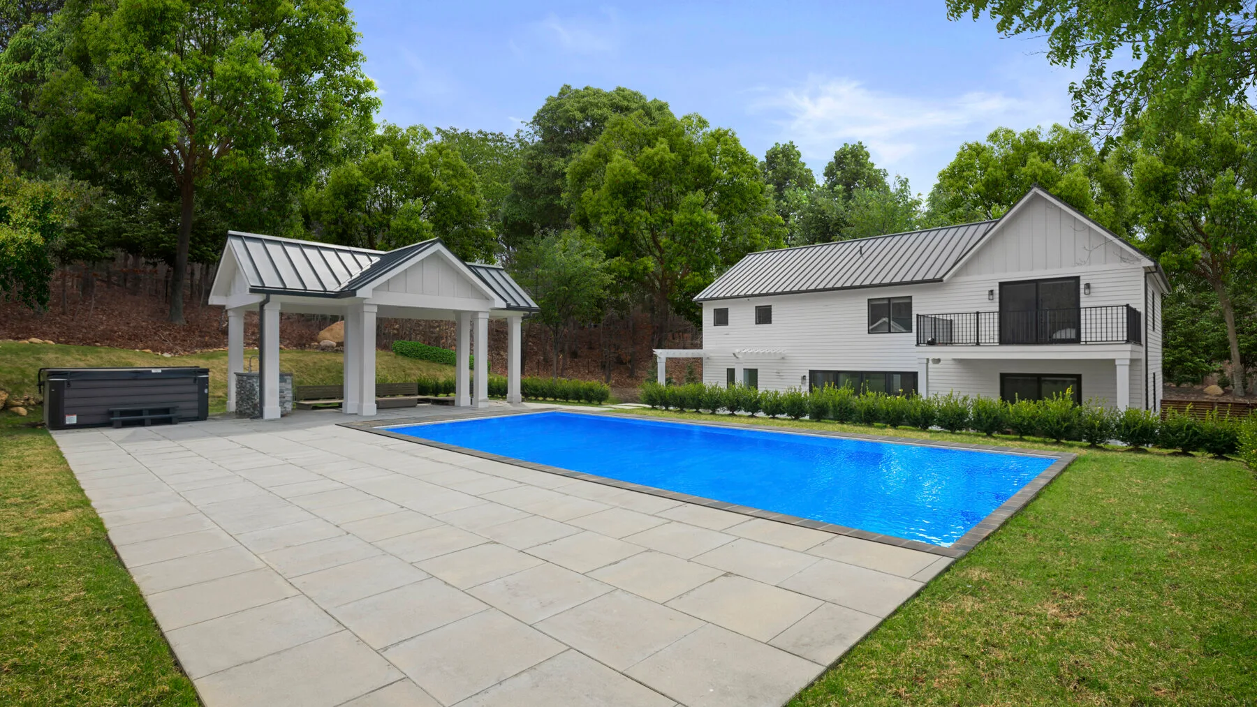 Una piscina y un patio pavimentado están junto a una casa blanca y árboles frondosos.