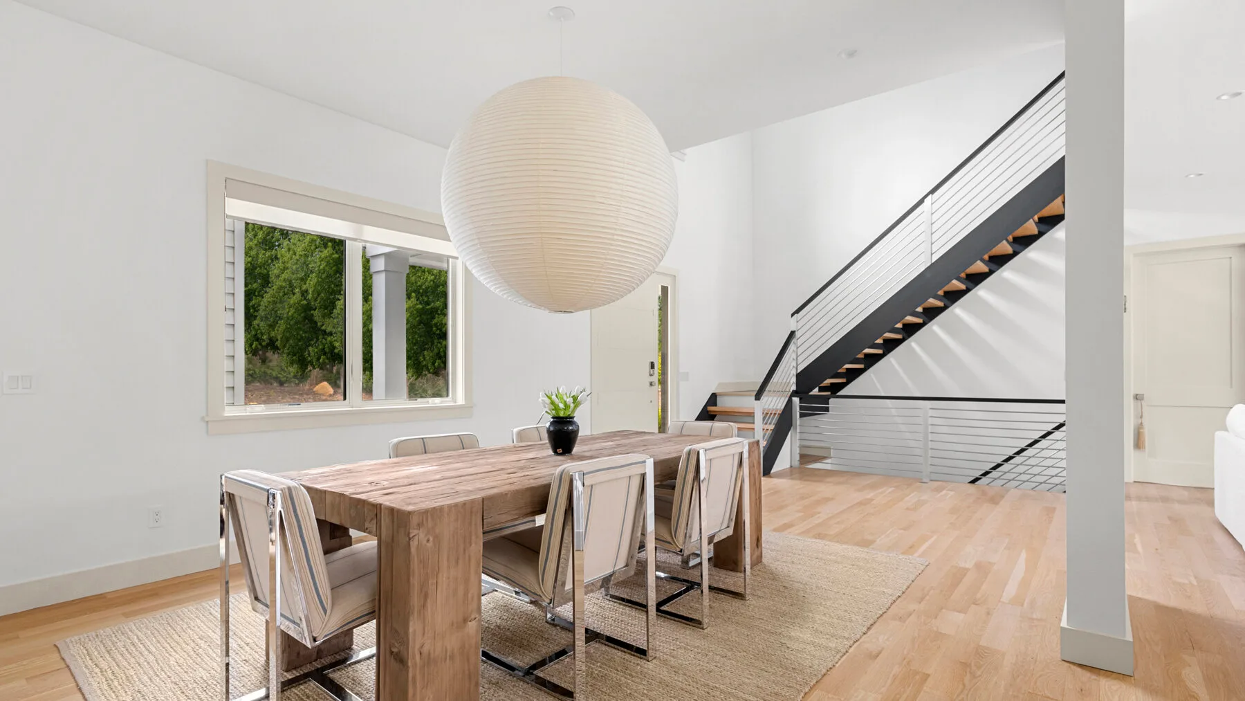Un comedor luminoso con una mesa de madera junto a una escalera y una ventana amplia.