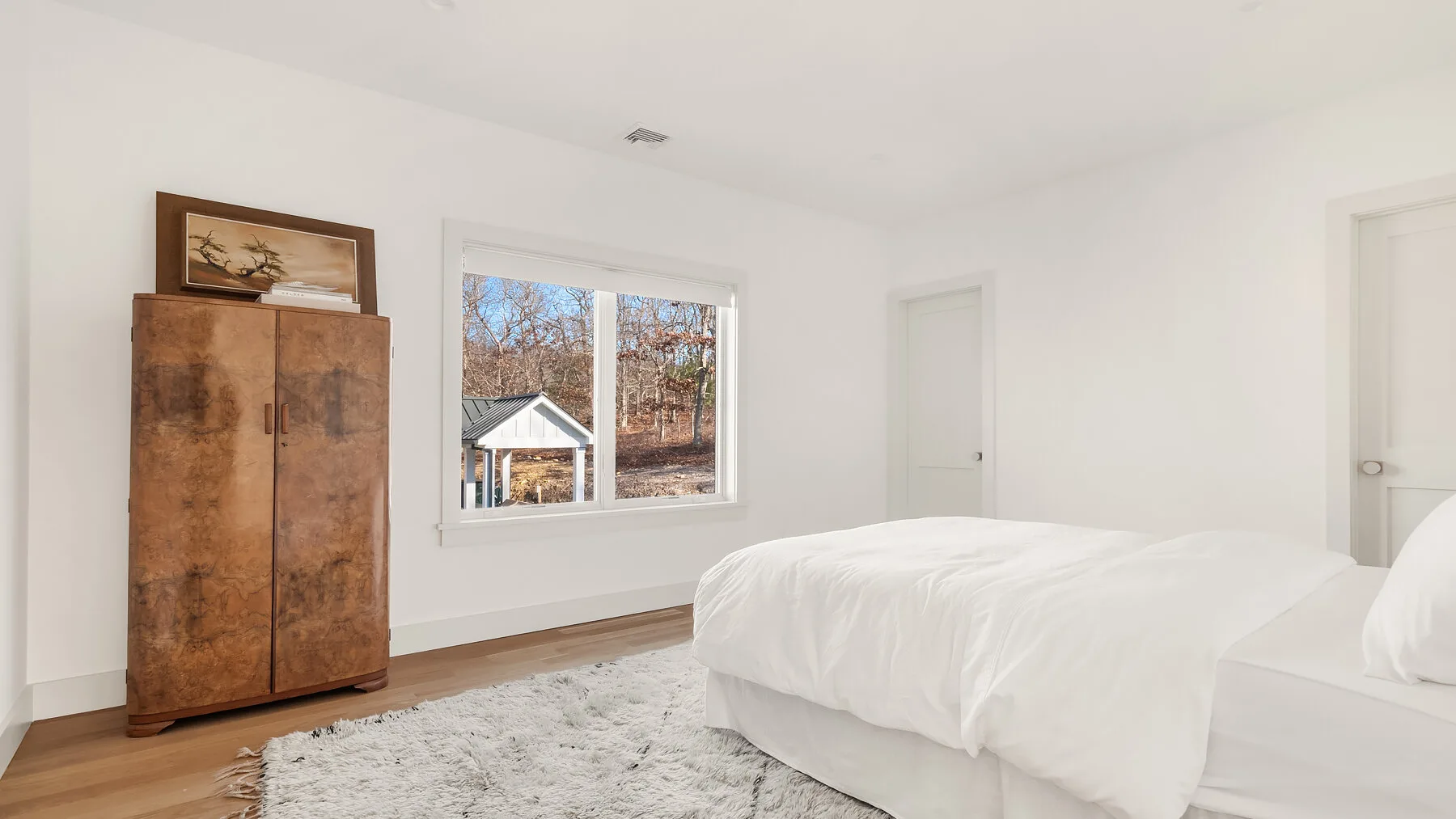 Un dormitorio luminoso con una cama blanca, un armario de madera y una ventana grande.