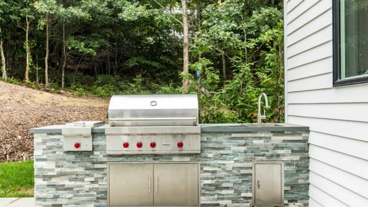 Una cocina exterior con parrilla empotrada y encimera revestida de piedra junto a una zona arbolada.