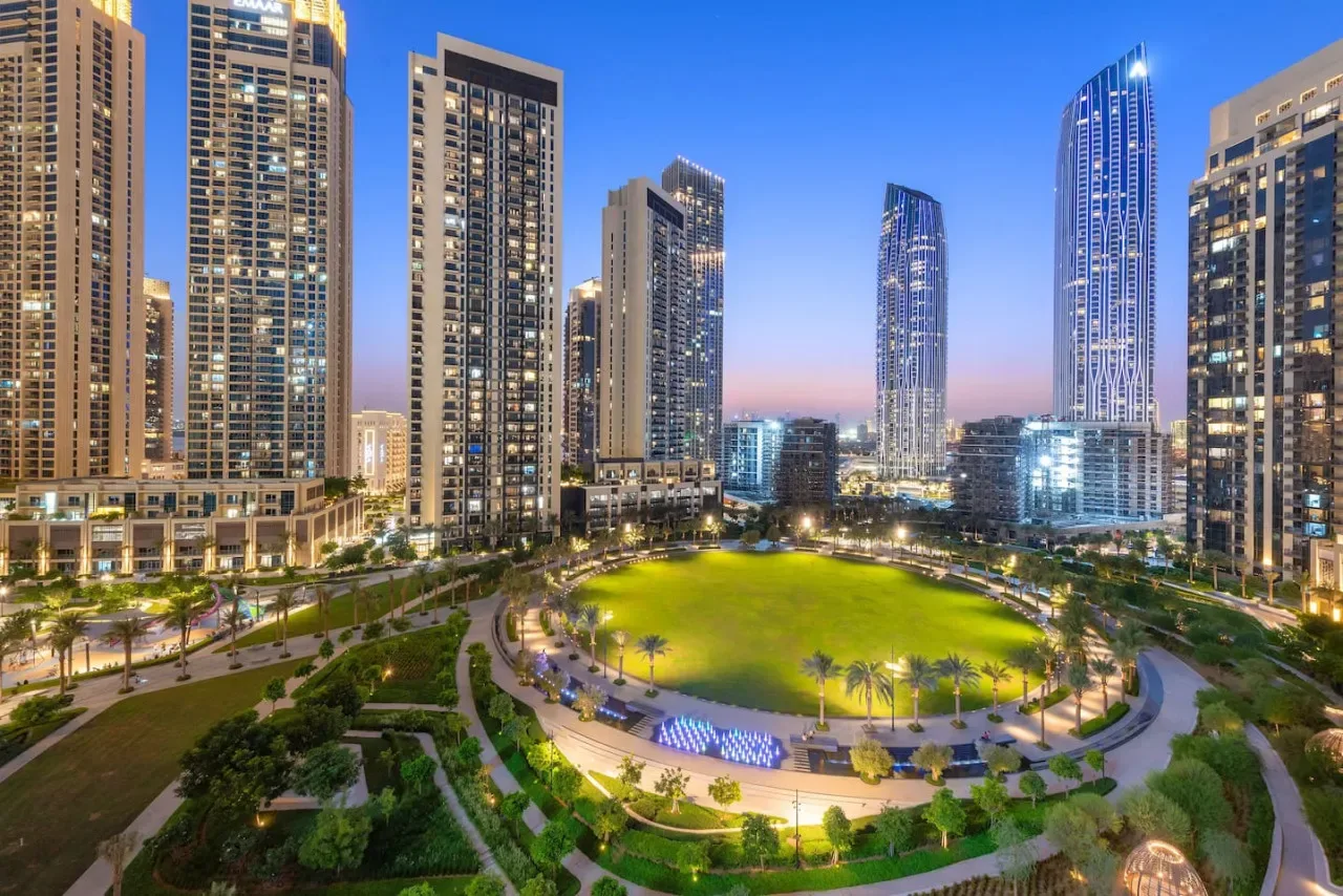 A landscaped city park with a broad green lawn, surrounded by illuminated high-rise buildings.