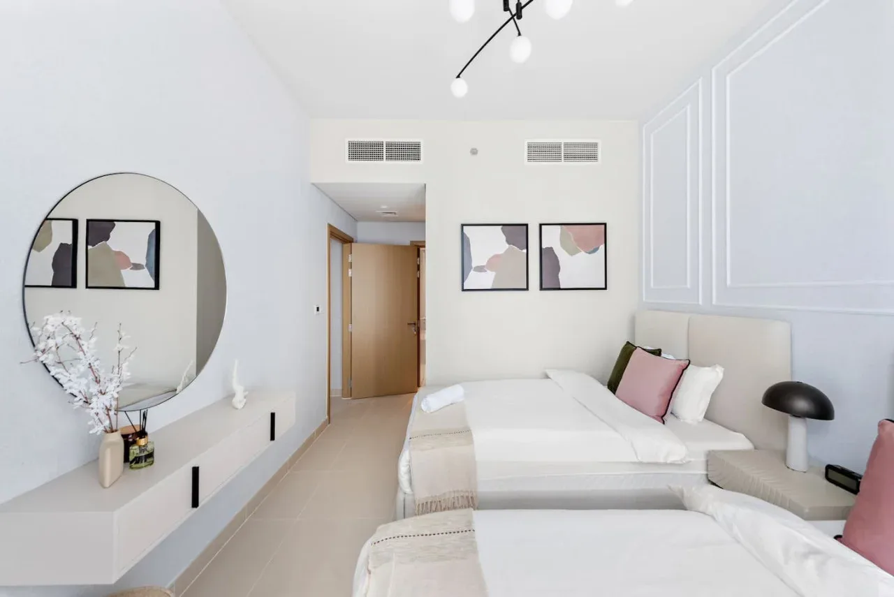 Un dormitorio luminoso con ropa de cama blanca, cuadros abstractos y un espejo redondo.
