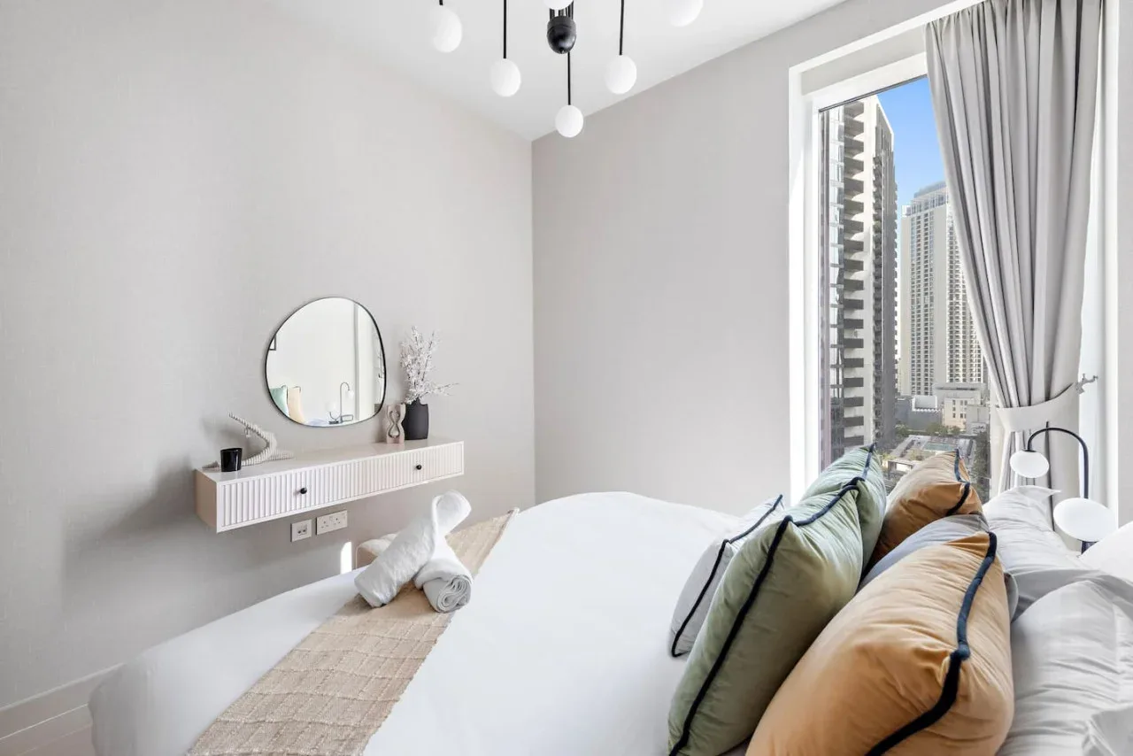 Un dormitorio luminoso con una cama junto a una ventana amplia con vista a edificios altos.