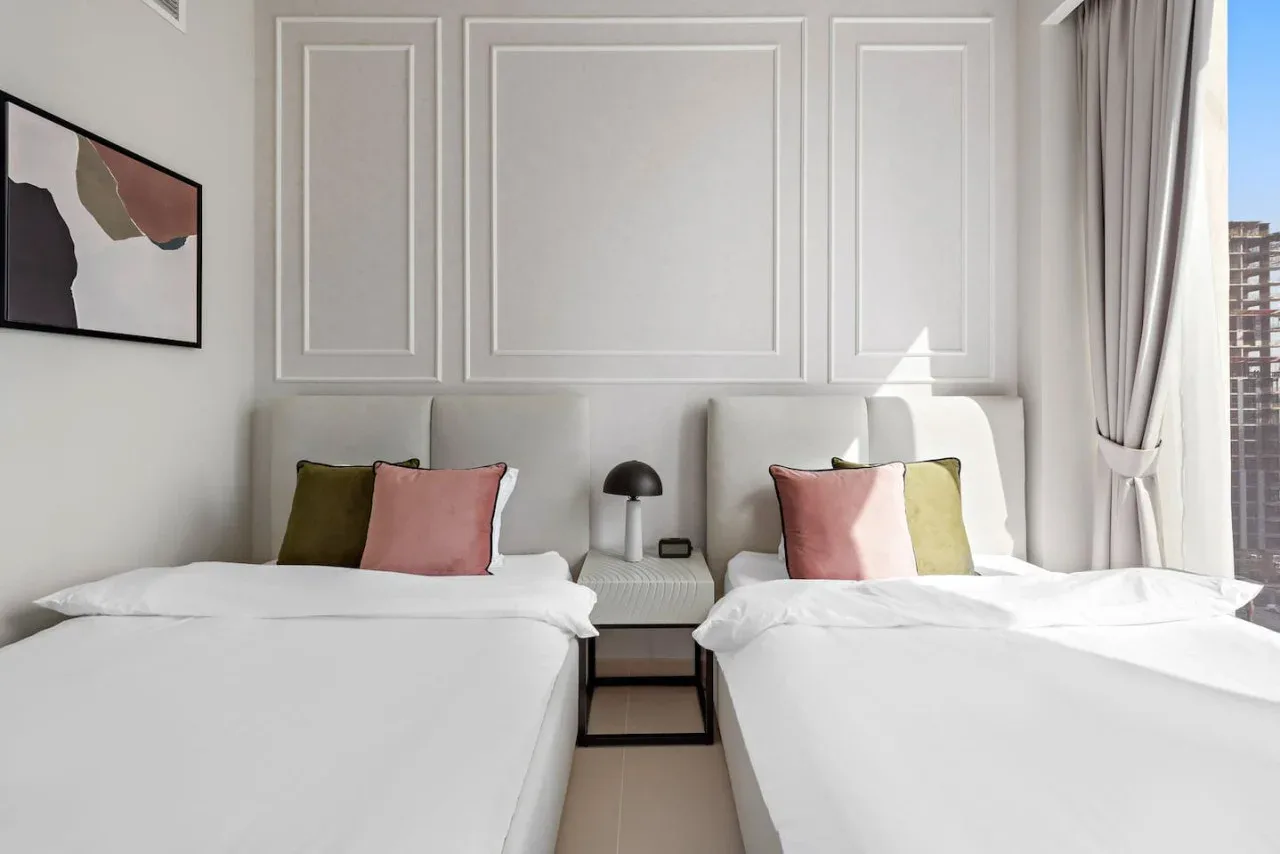 Un dormitorio luminoso con dos camas, cojines de colores y una pequeña mesita de noche.