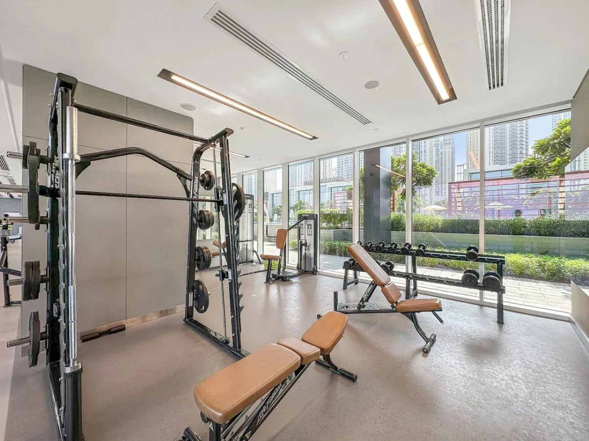 Un gimnasio con máquinas de pesas y bancos junto a grandes ventanales.