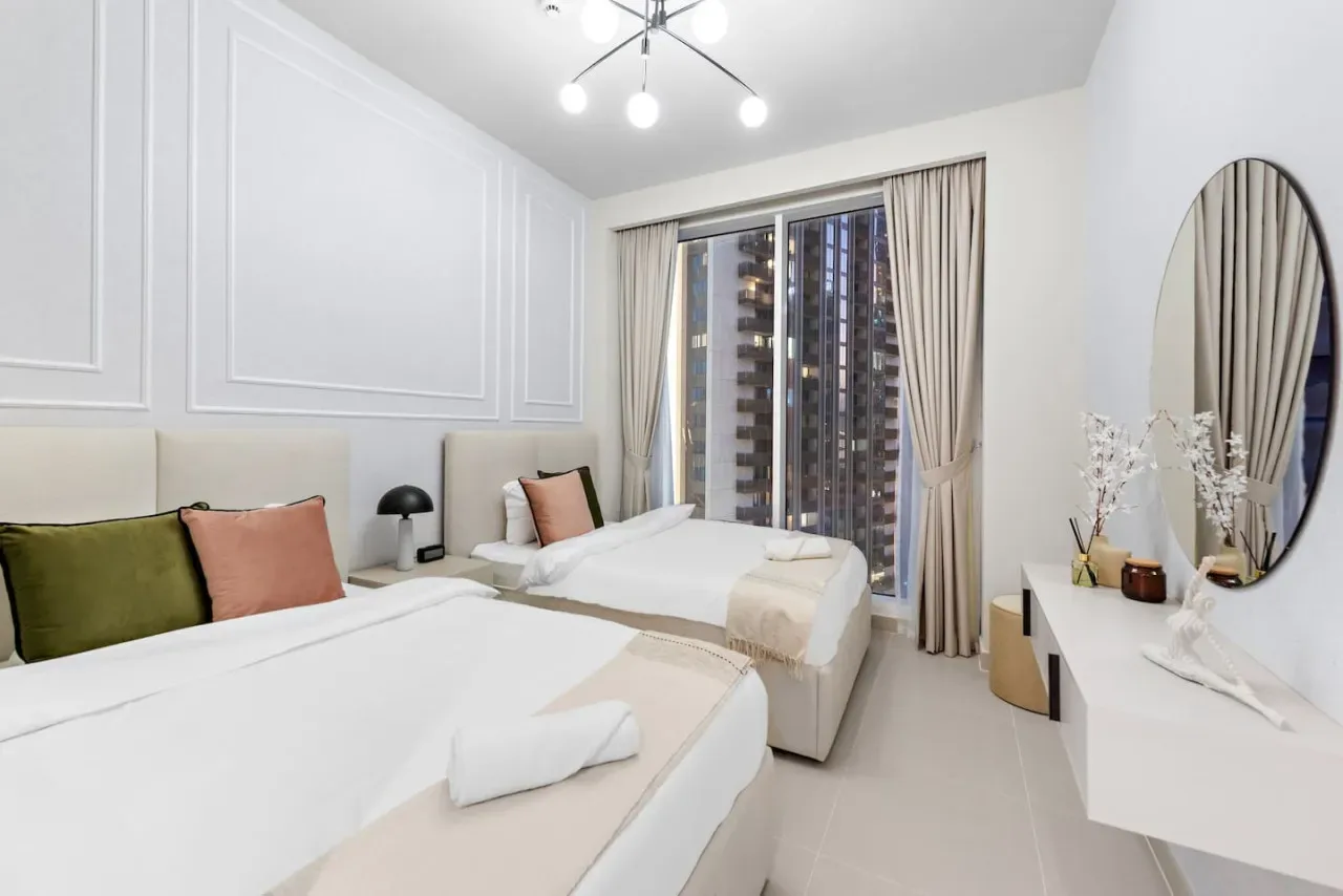A bright bedroom with two beds and a large window overlooking nearby buildings.