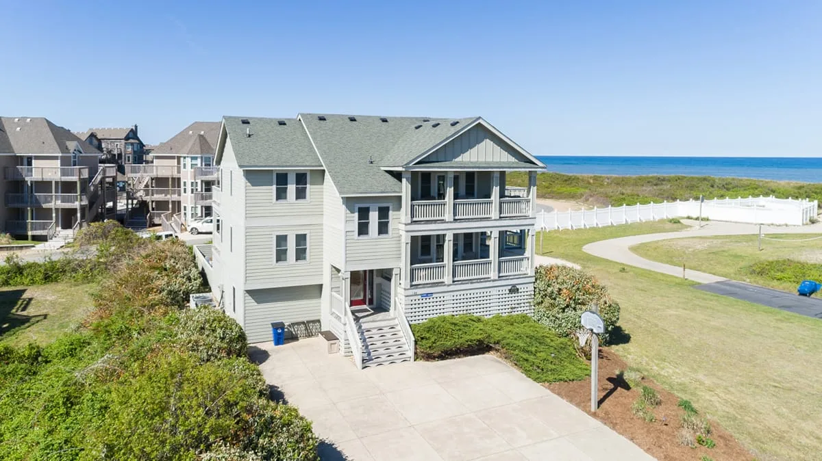A large coastal house stands beside a grassy field, with the ocean visible beyond.