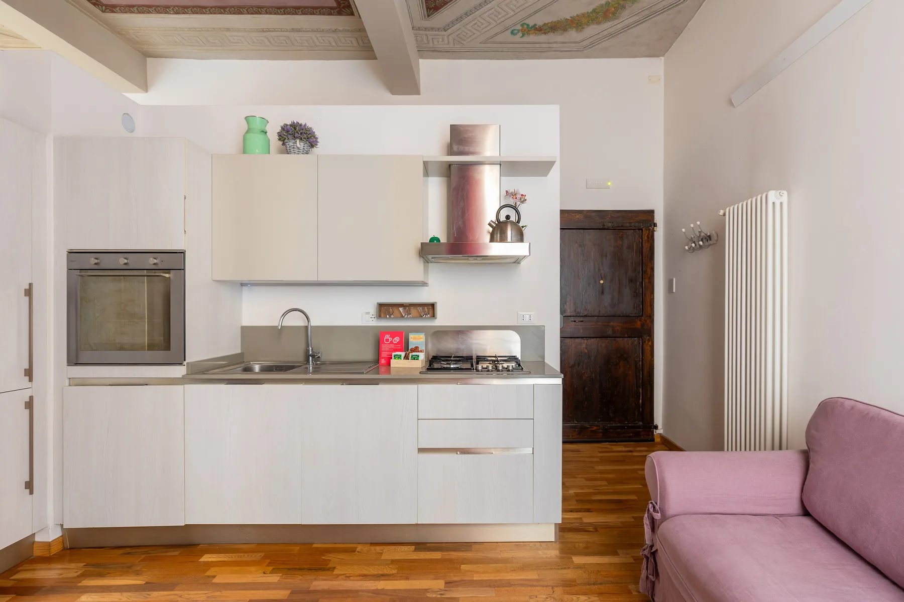 Una cocina compacta con gabinetes blancos, una placa de cocina y un sofá rosa cerca.