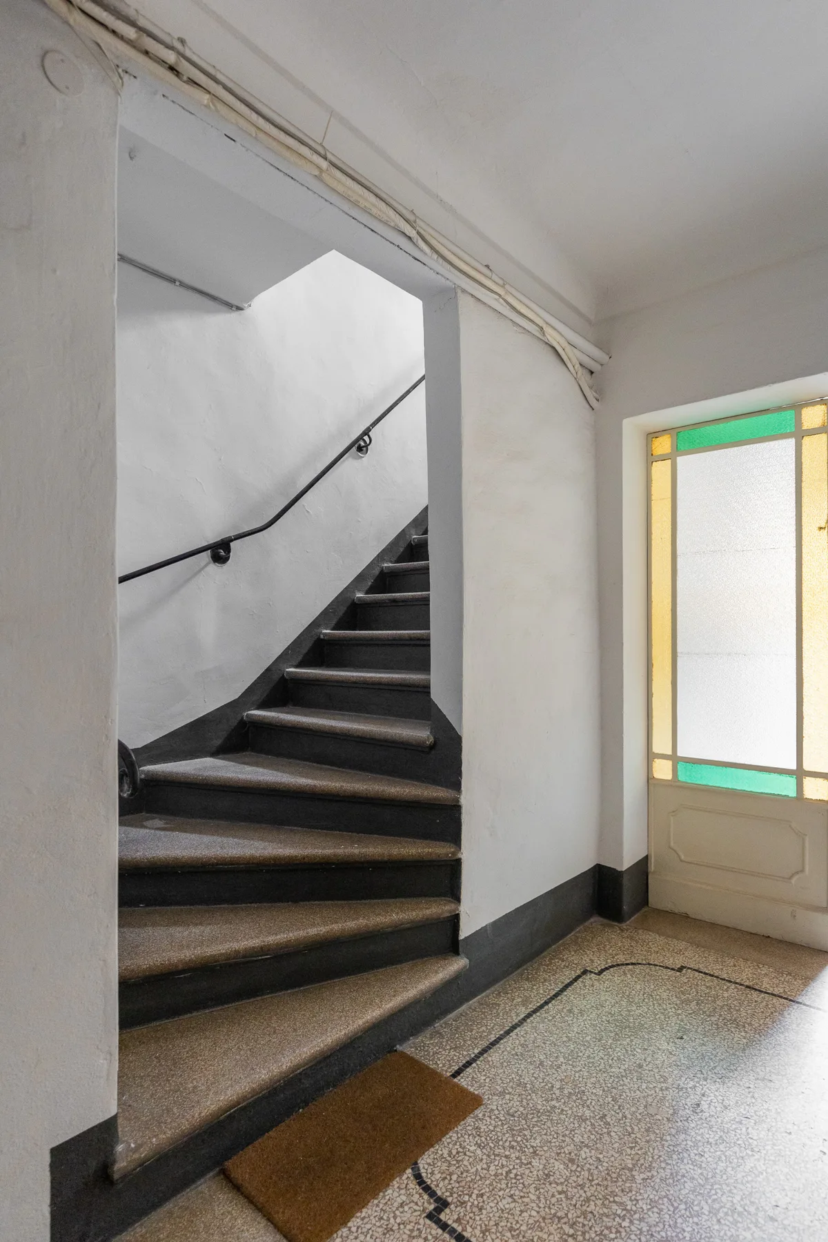Una escalera interior con peldaños oscuros junto a una puerta de vidrio de colores.