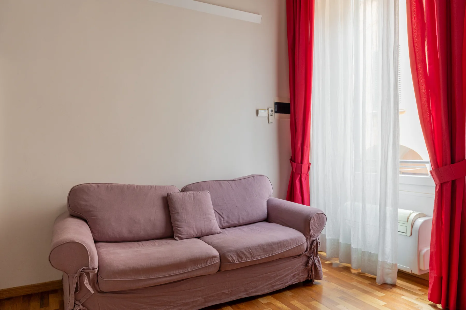 Una sala de estar con asientos color malva junto a una ventana con cortinas translúcidas y rojas.