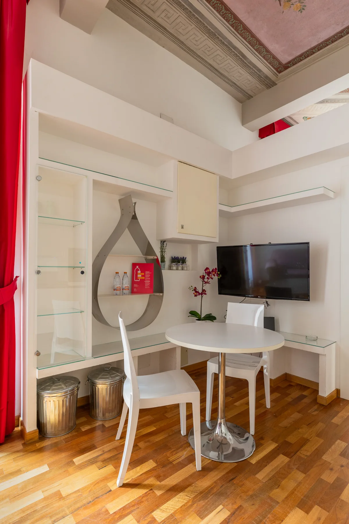 Una luminosa zona de estar tiene una mesa redonda, asientos blancos y un televisor en la pared.