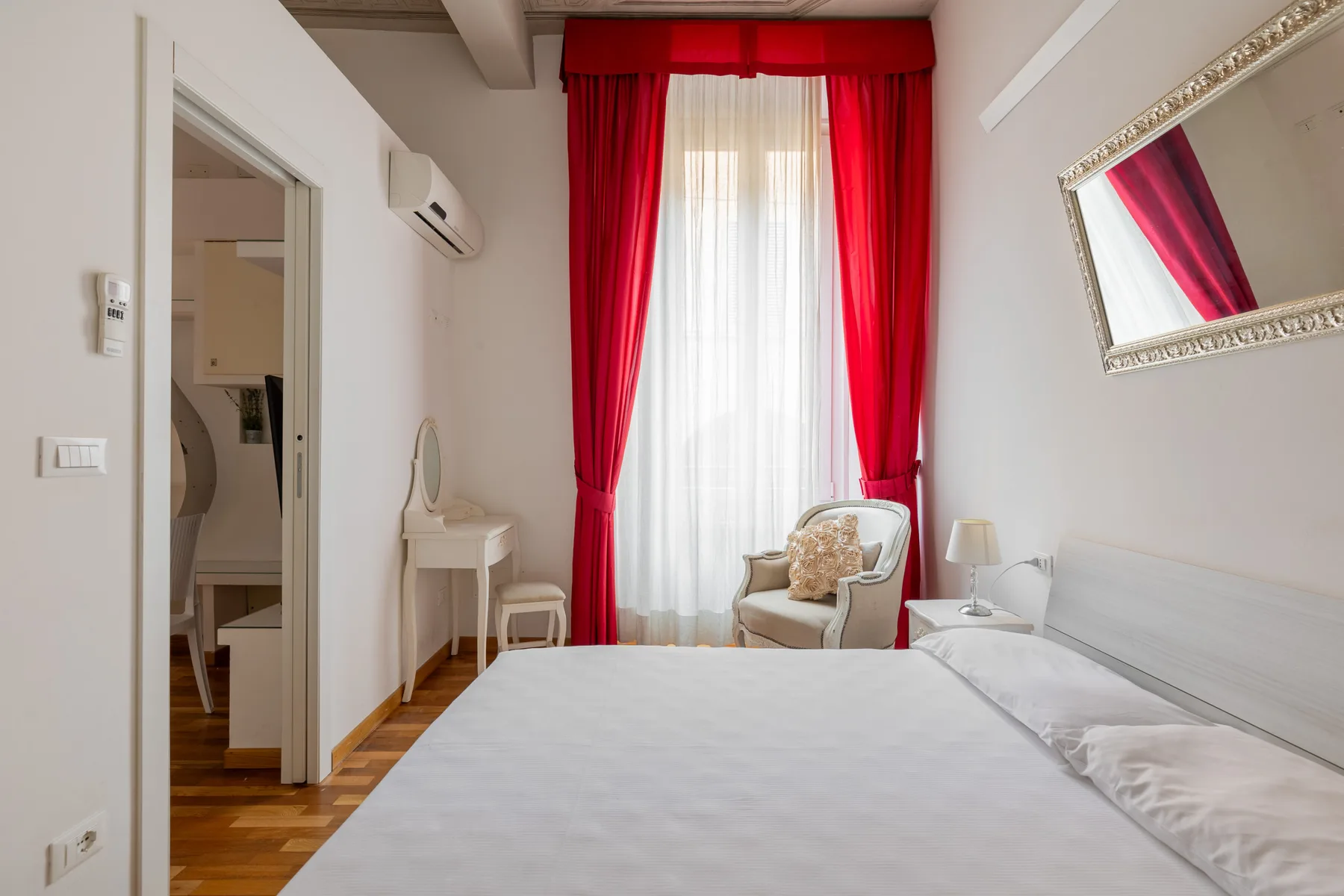 Un dormitorio luminoso con una cama grande, cortinas rojas y una pequeña zona de estar.