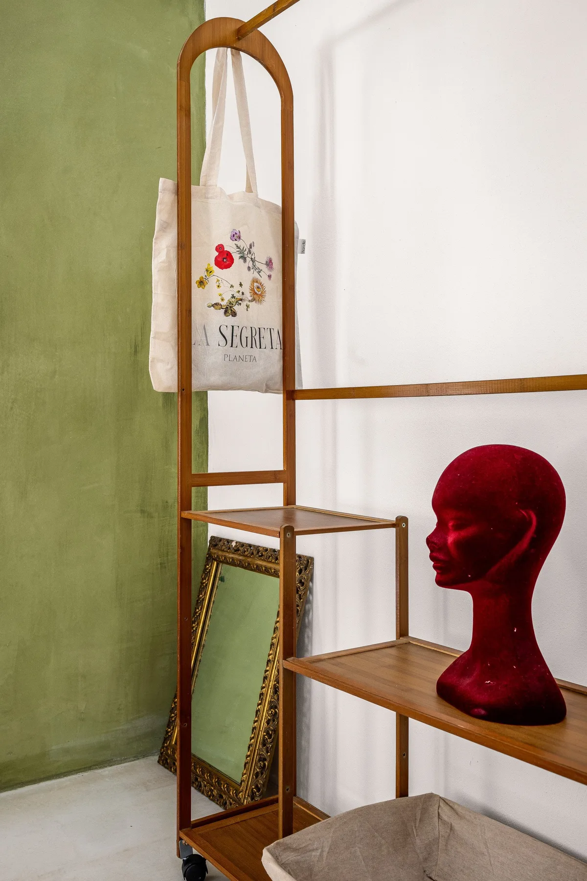 Una estantería de exposición está junto a una pared blanca y otra verde, con un busto rojo y un espejo enmarcado.