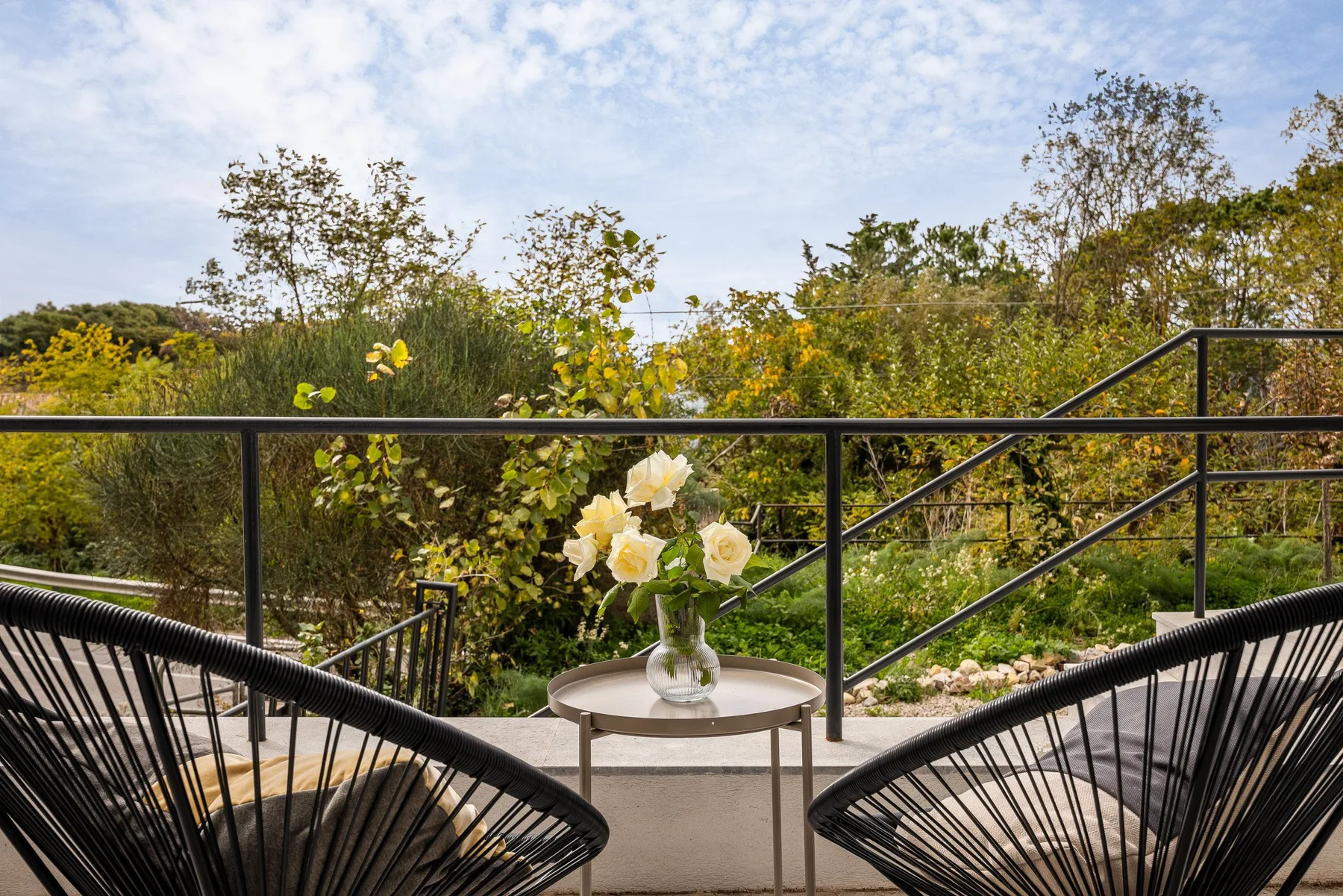 Un patio al aire libre con asientos y una mesita con flores da a una zona verde.